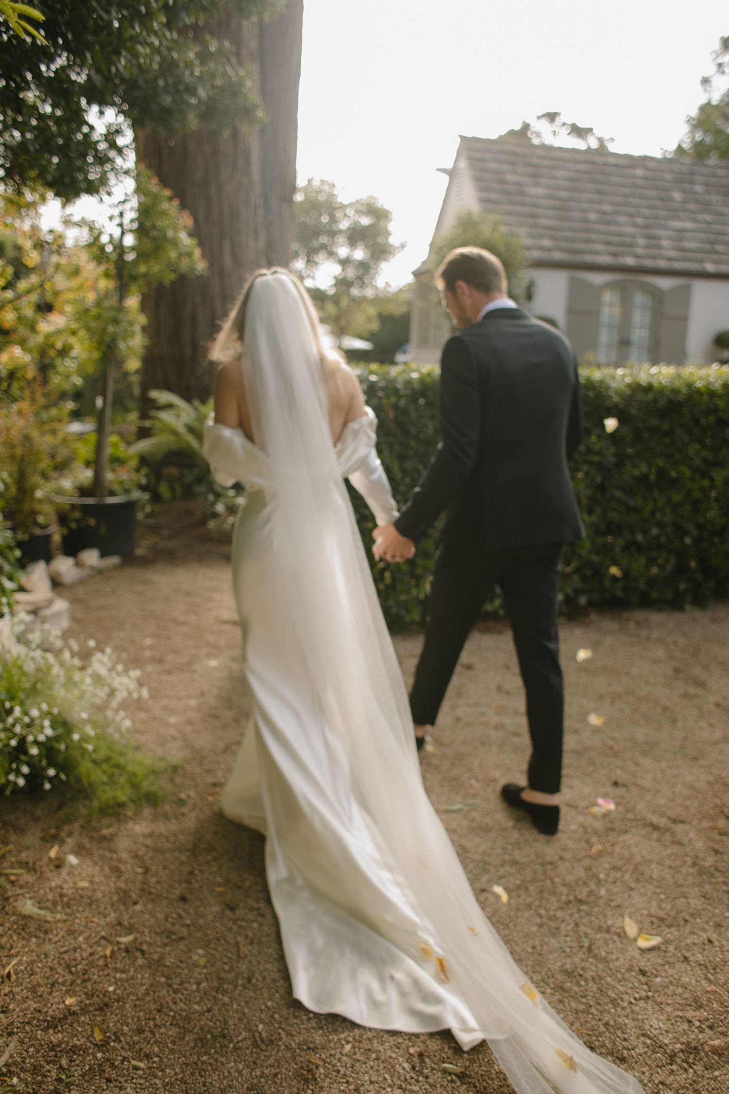 A wedding couple walks along a garden path with a chapel in the background as the bride's flowing white dress trails behind.