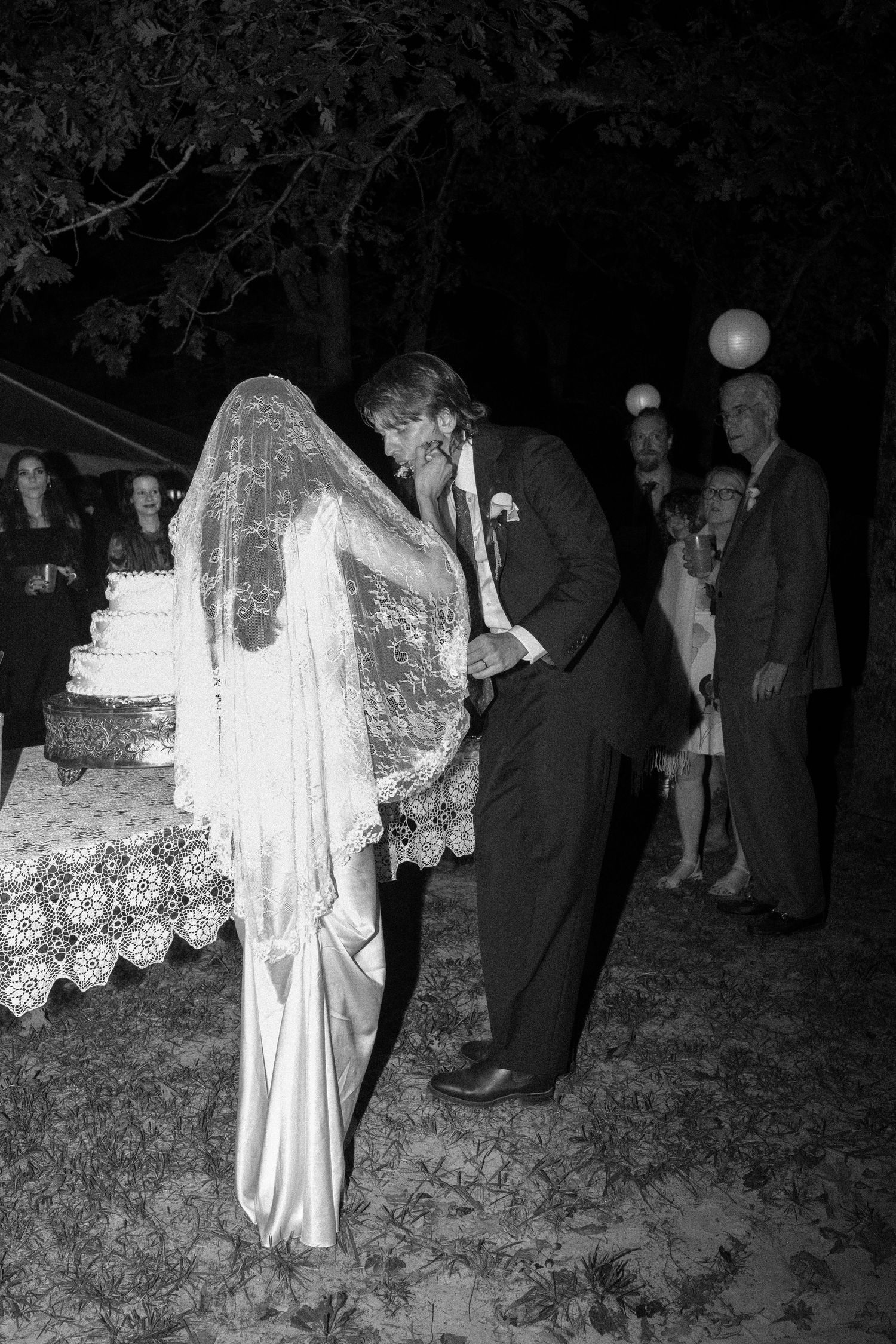 Black and white photo of bride and groom cutting wedding cake outdoors at night with guests watching.