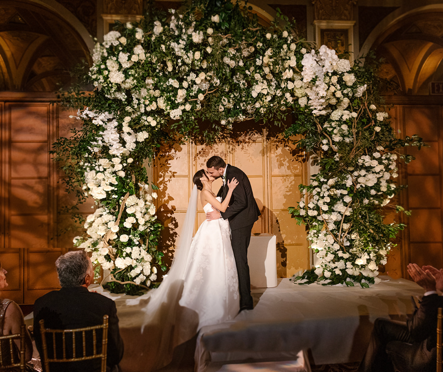 A wedding ceremony moment captured under a lush white floral arch in an elegant indoor venue.