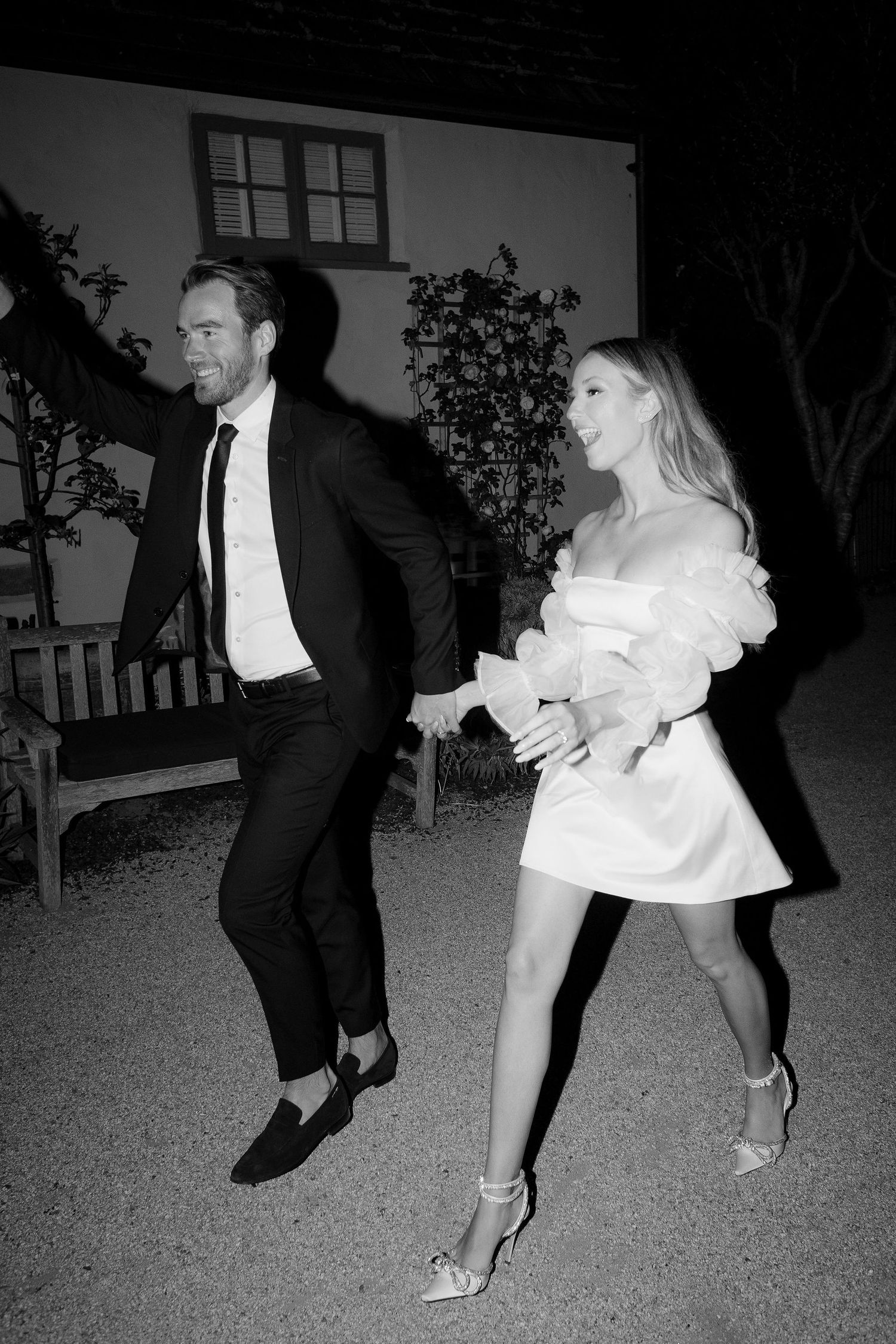 Couple in formal attire dancing outdoors at night, man in black suit, woman in white mini dress with heels.