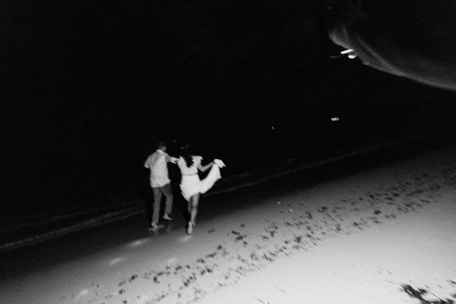 Black and white image of two figures strolling along a dark beach leaving footprints in the sand.