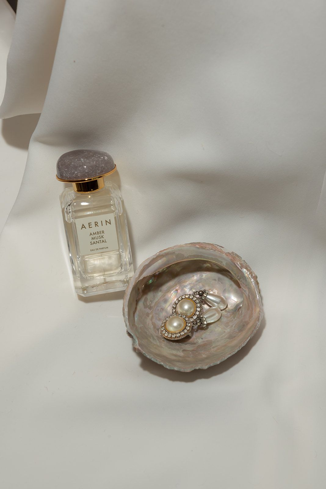 A bottle of Aerin perfume and jewelry displayed on a pearly shell dish against a white background.