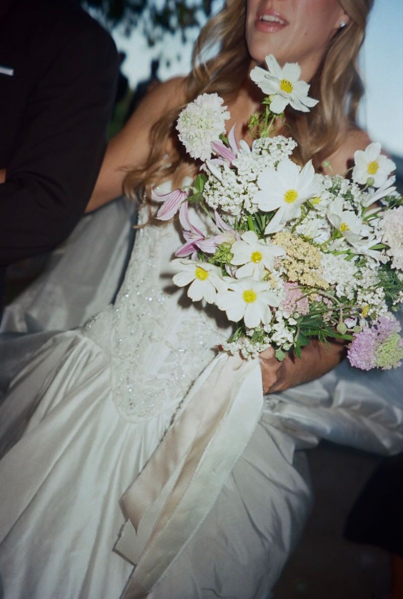 A wedding bouquet of white daisies and lilac flowers rests against a flowing white dress in a series of closeup shots.