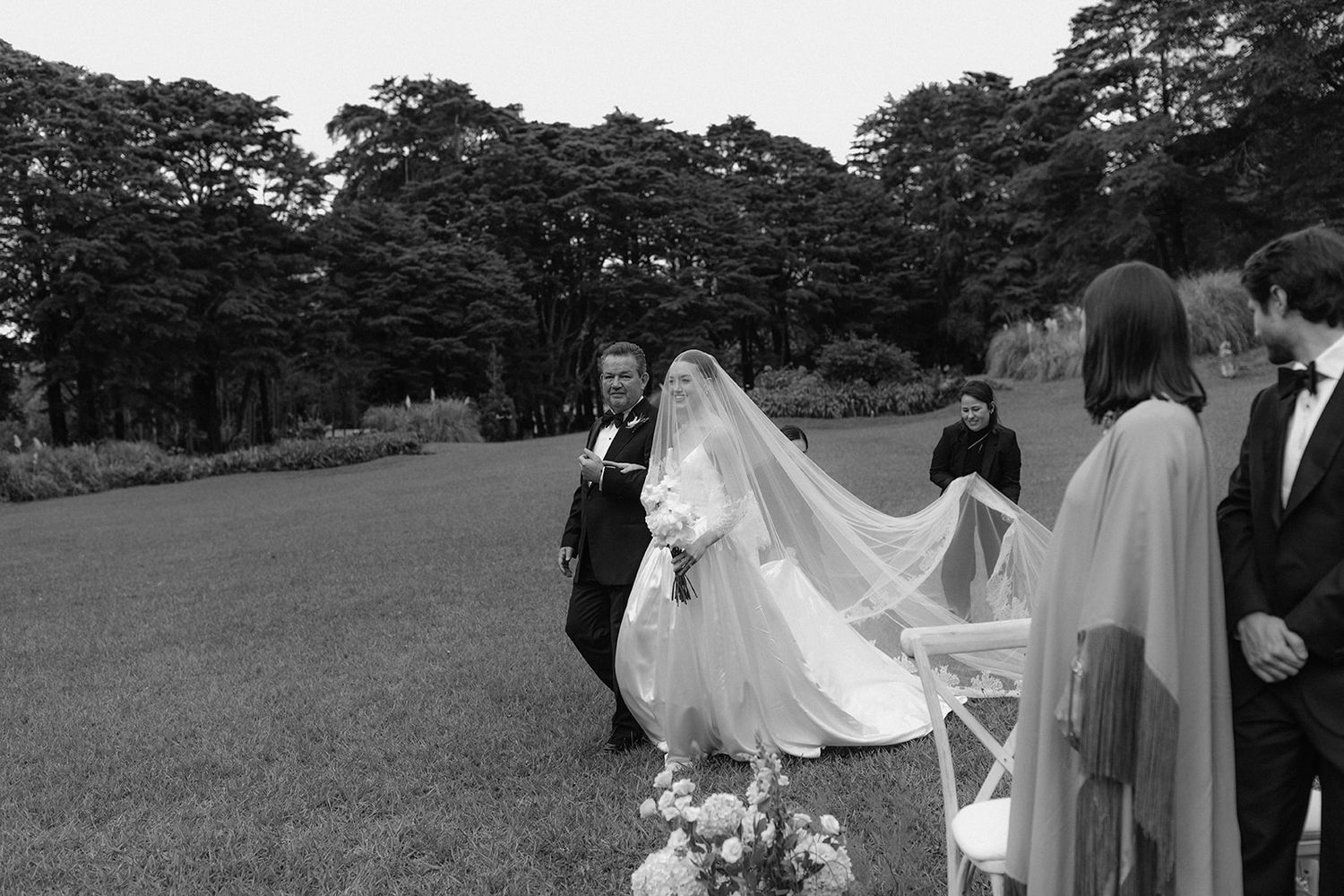 A wedding party walks through a tree-lined path during an outdoor celebration in black and white photography.