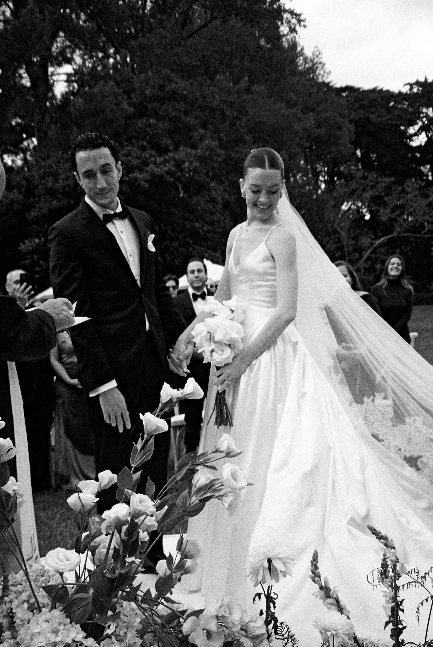 A wedding couple poses joyfully outdoors in black and white as the bride's flowing veil catches the breeze.