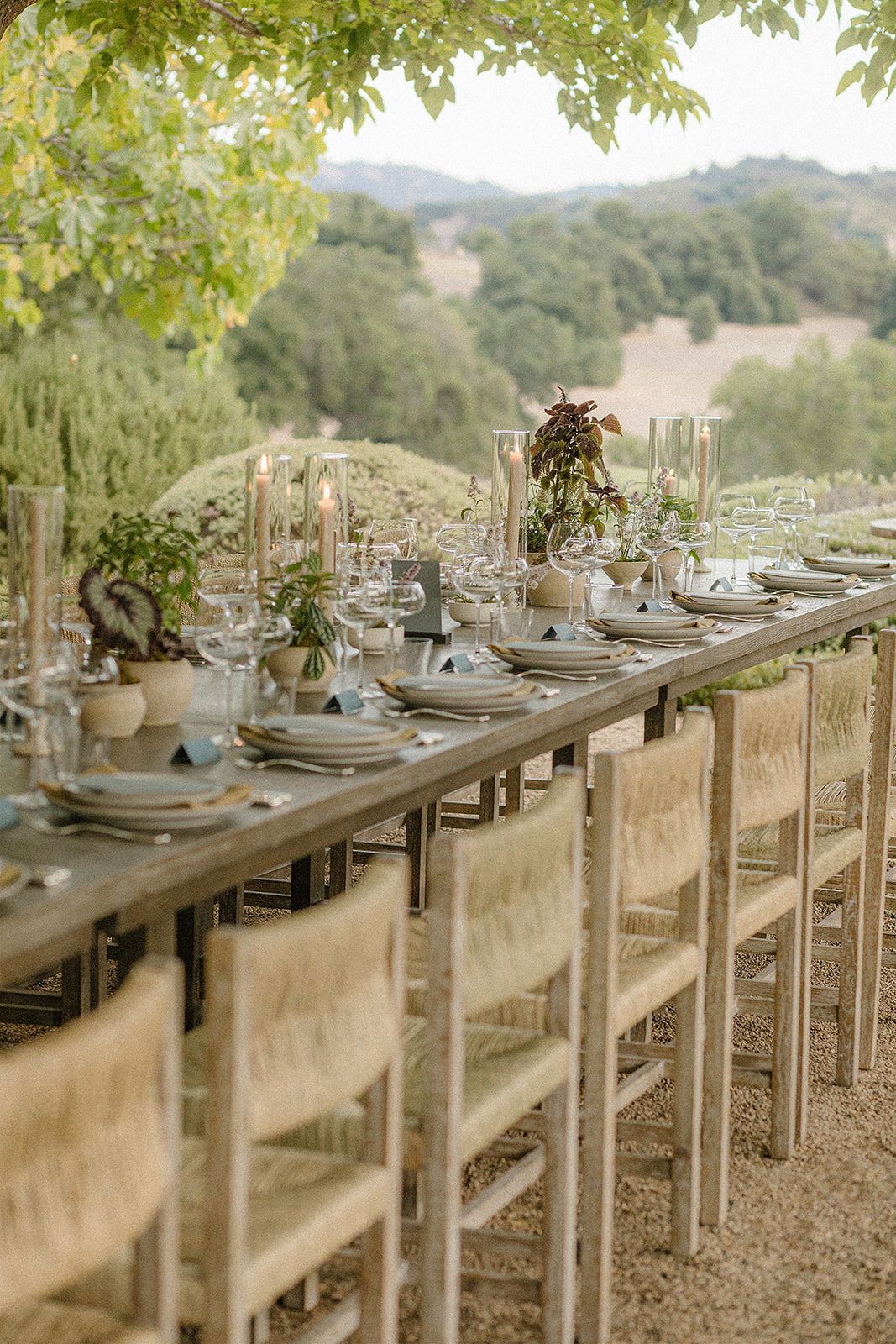 Elegant outdoor dining table set for a wedding reception with rustic chairs, candles, and greenery centerpieces in a scenic hillside.