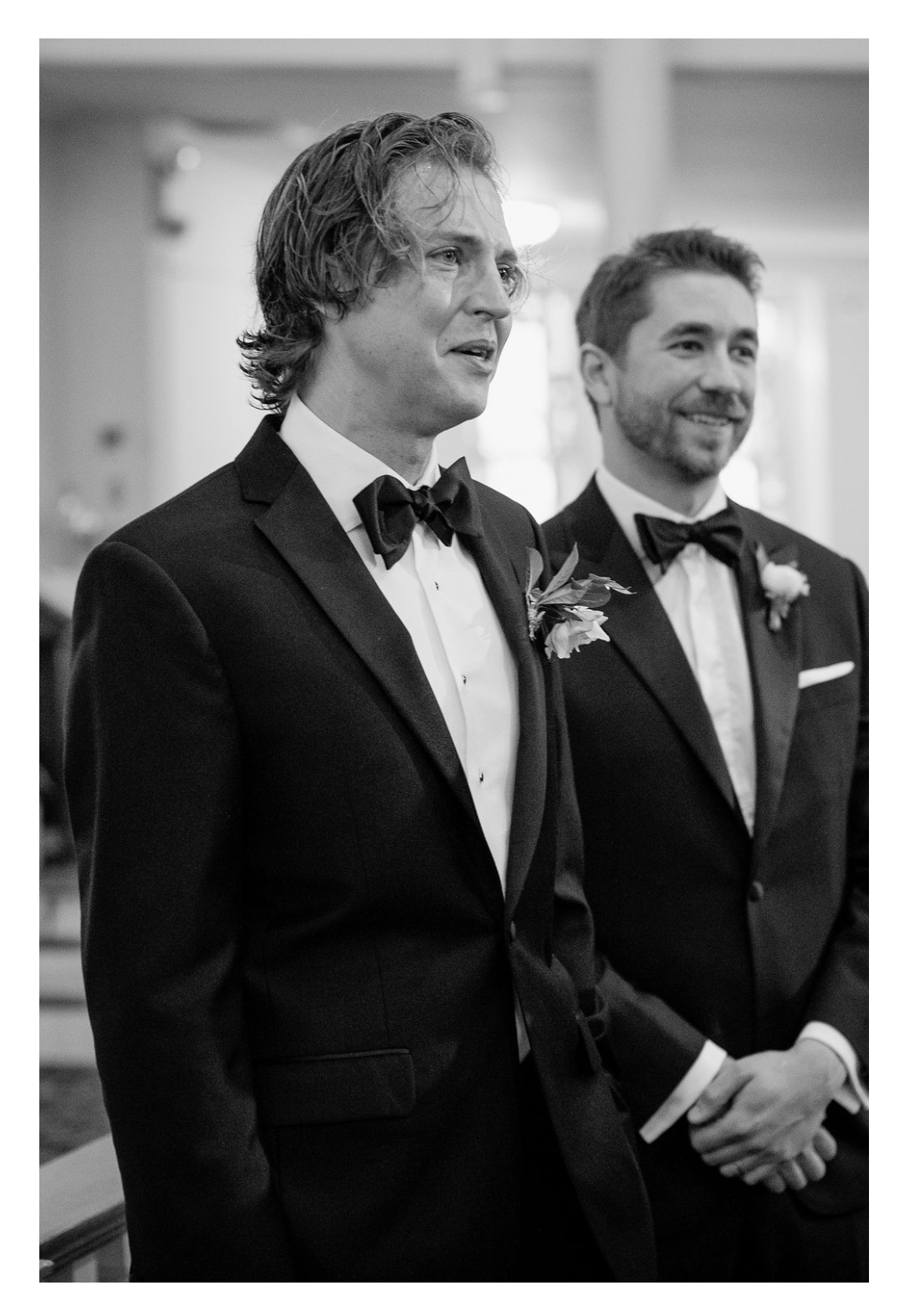 Two gentlemen in black tuxedos and bow ties share a candid moment of laughter at a formal event.