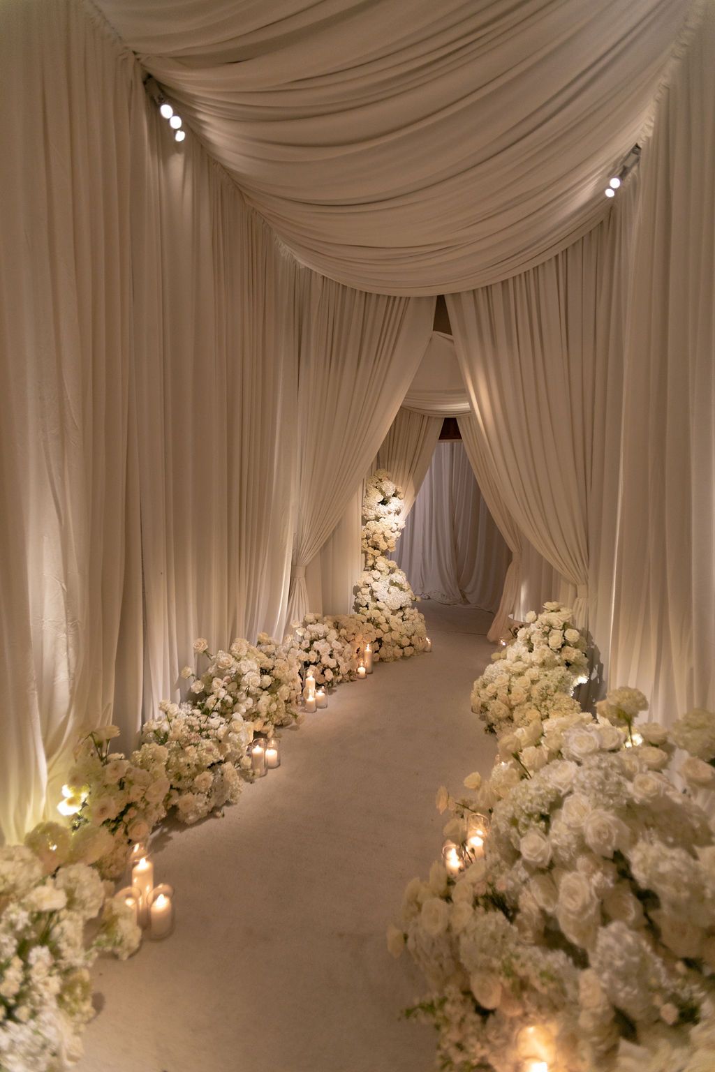 Elegant wedding aisle with white draping, floral arrangements, and candlelight creating a romantic ceremonial entrance.