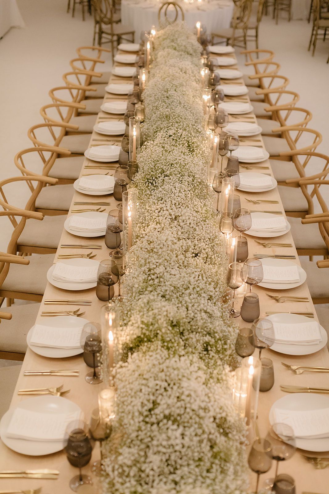 Elegant wedding reception table with baby's breath floral runner, white plates, gold cutlery, and glowing candles on a long banquet table.