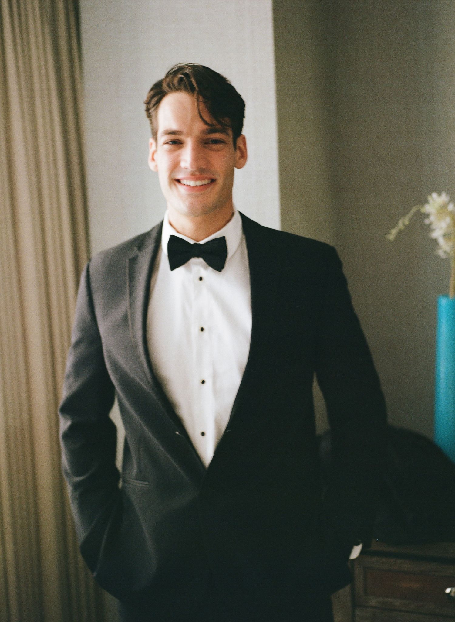 A groom in a classic black tuxedo and white bow tie smiles while getting ready for his wedding day.