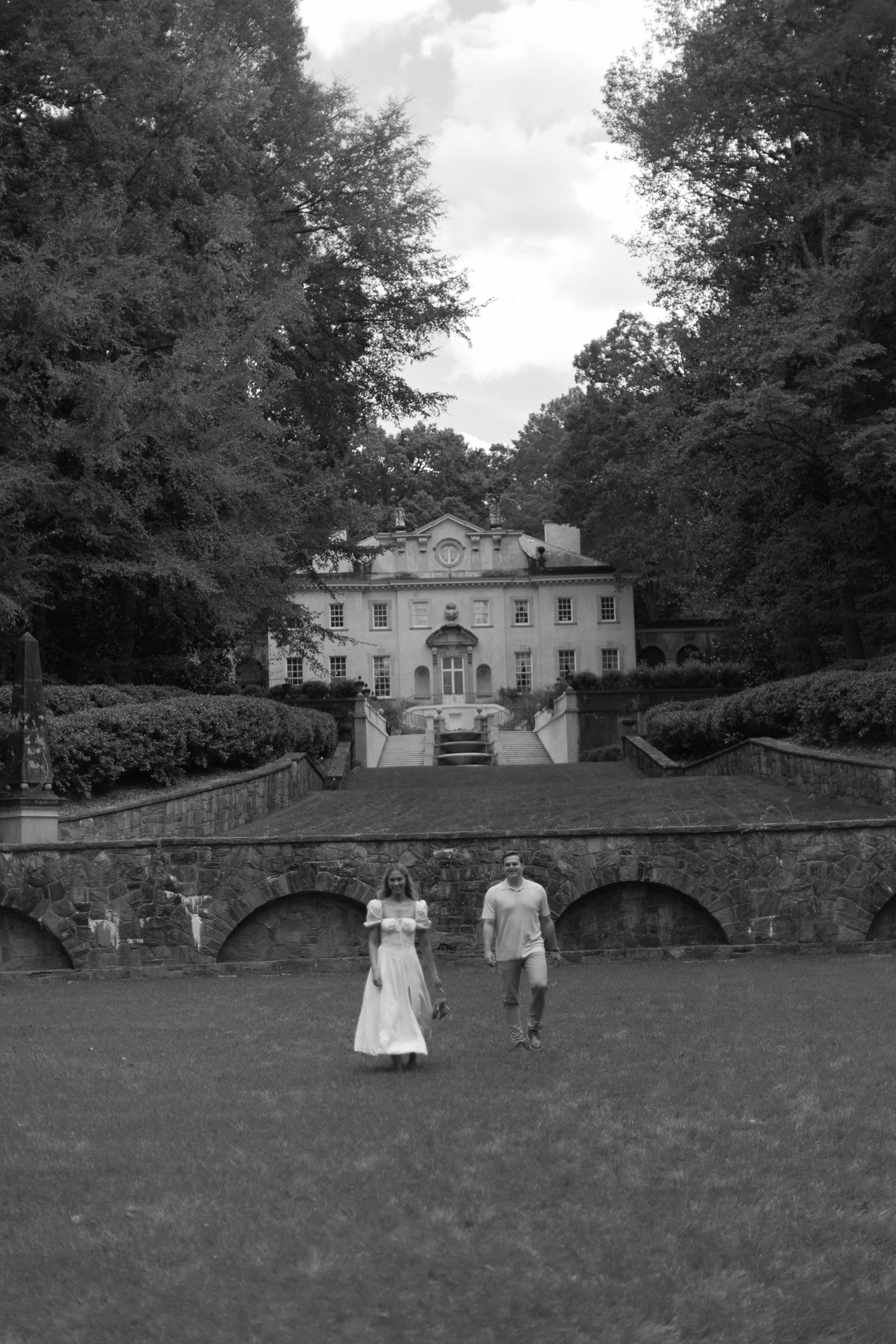 Historic black and white photo showing people walking on a lawn near a grand estate house with stone bridge and trees.