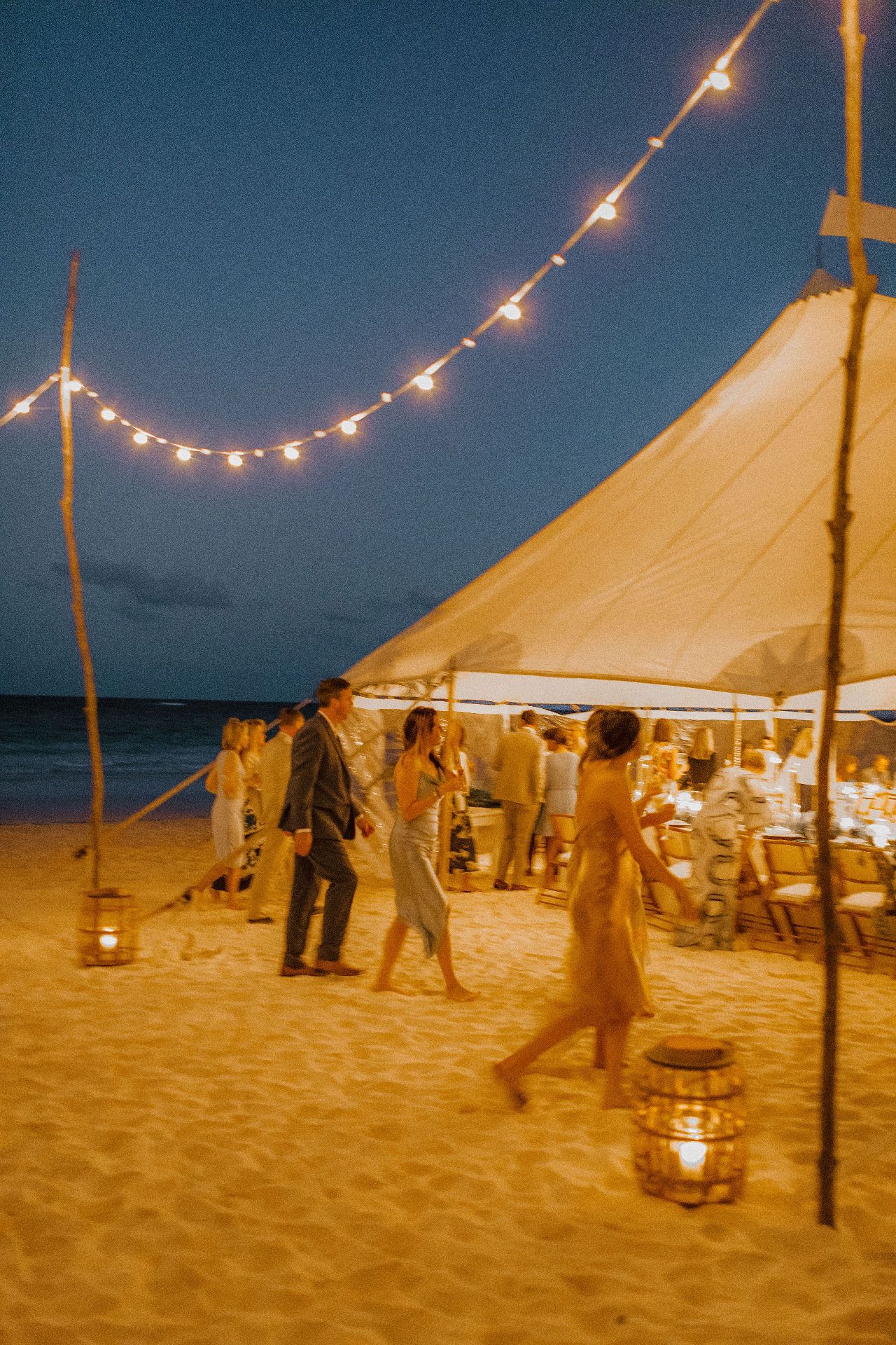String lights illuminate a beach tent party at dusk with people mingling on the sand.