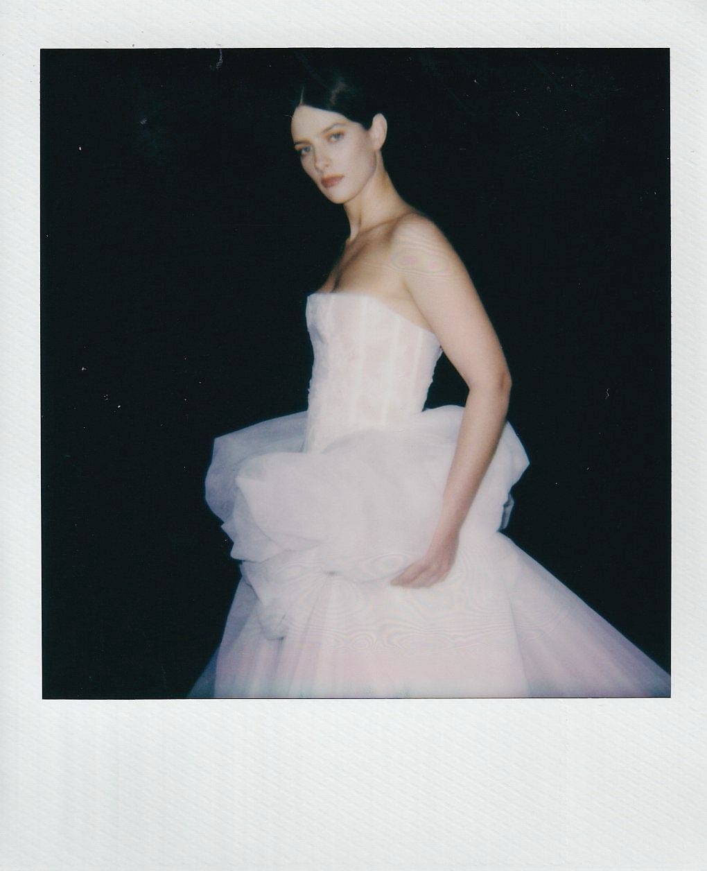 A figure in an elegant white strapless tulle wedding gown poses against a dark background in a Polaroid-style photo.