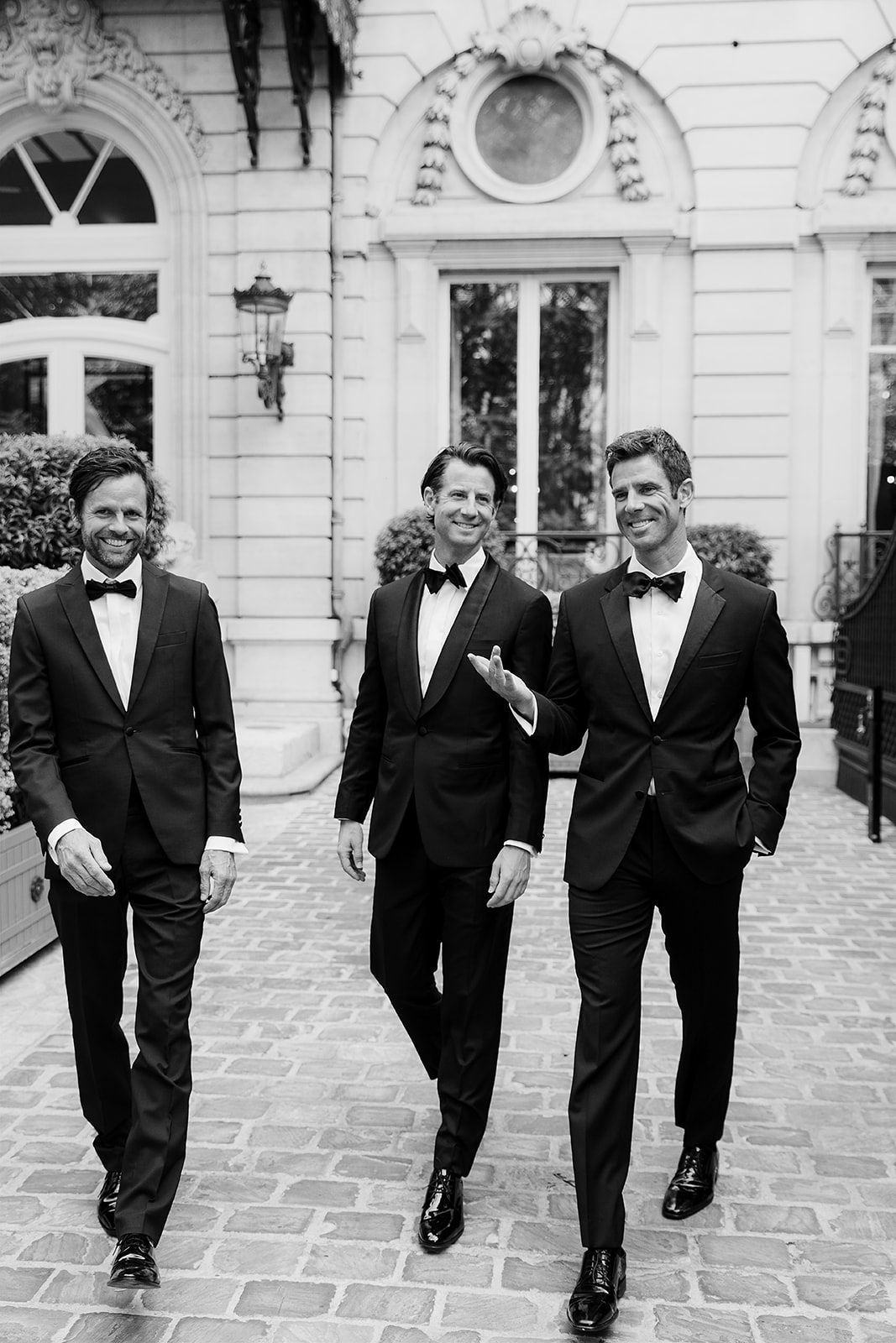 Three groomsmen in black tuxedos and bow ties walking together outside an elegant building in a black and white photo.