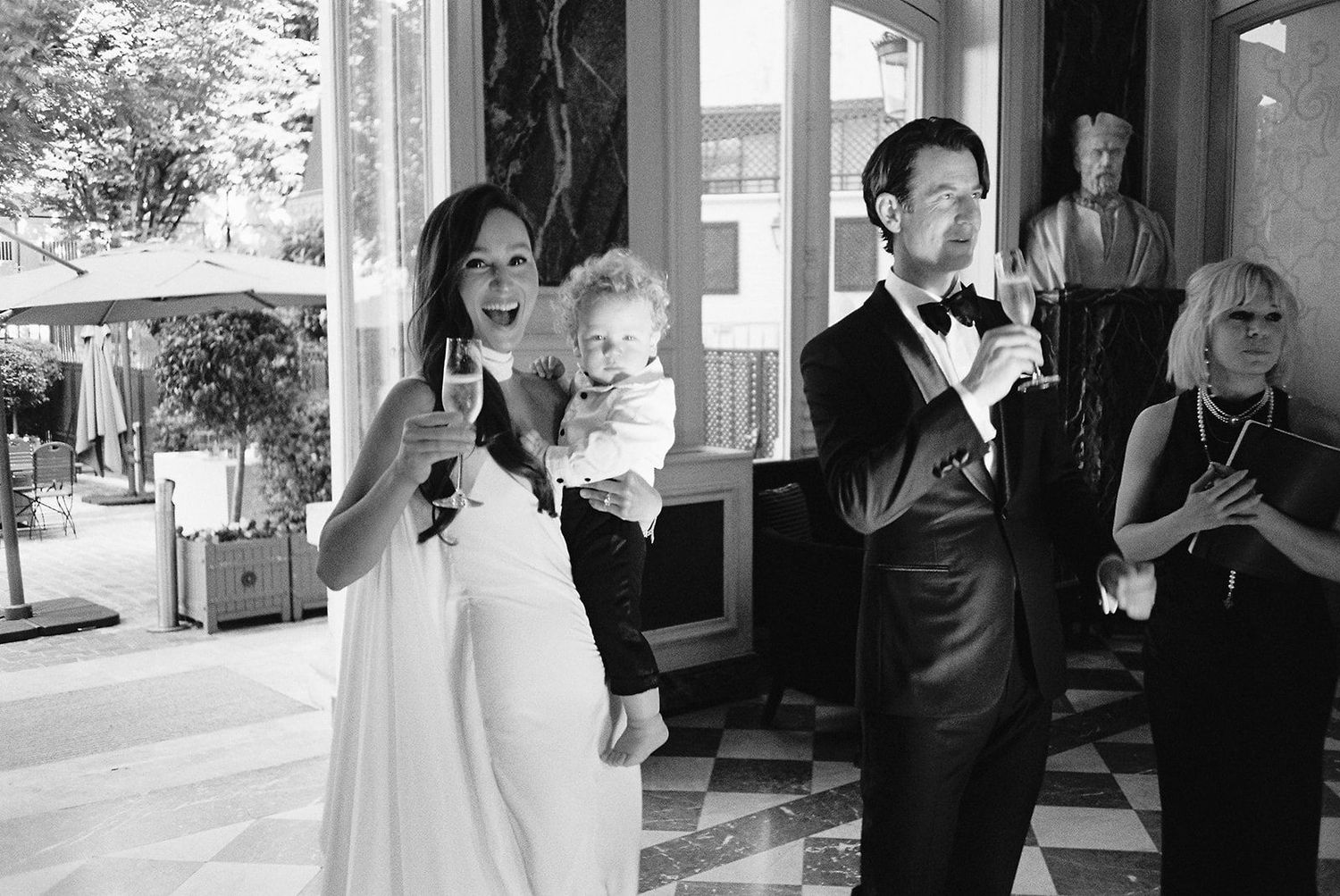 Black and white photo of a bride holding a toddler and a groom in a tuxedo raising glasses for a toast at a wedding reception.