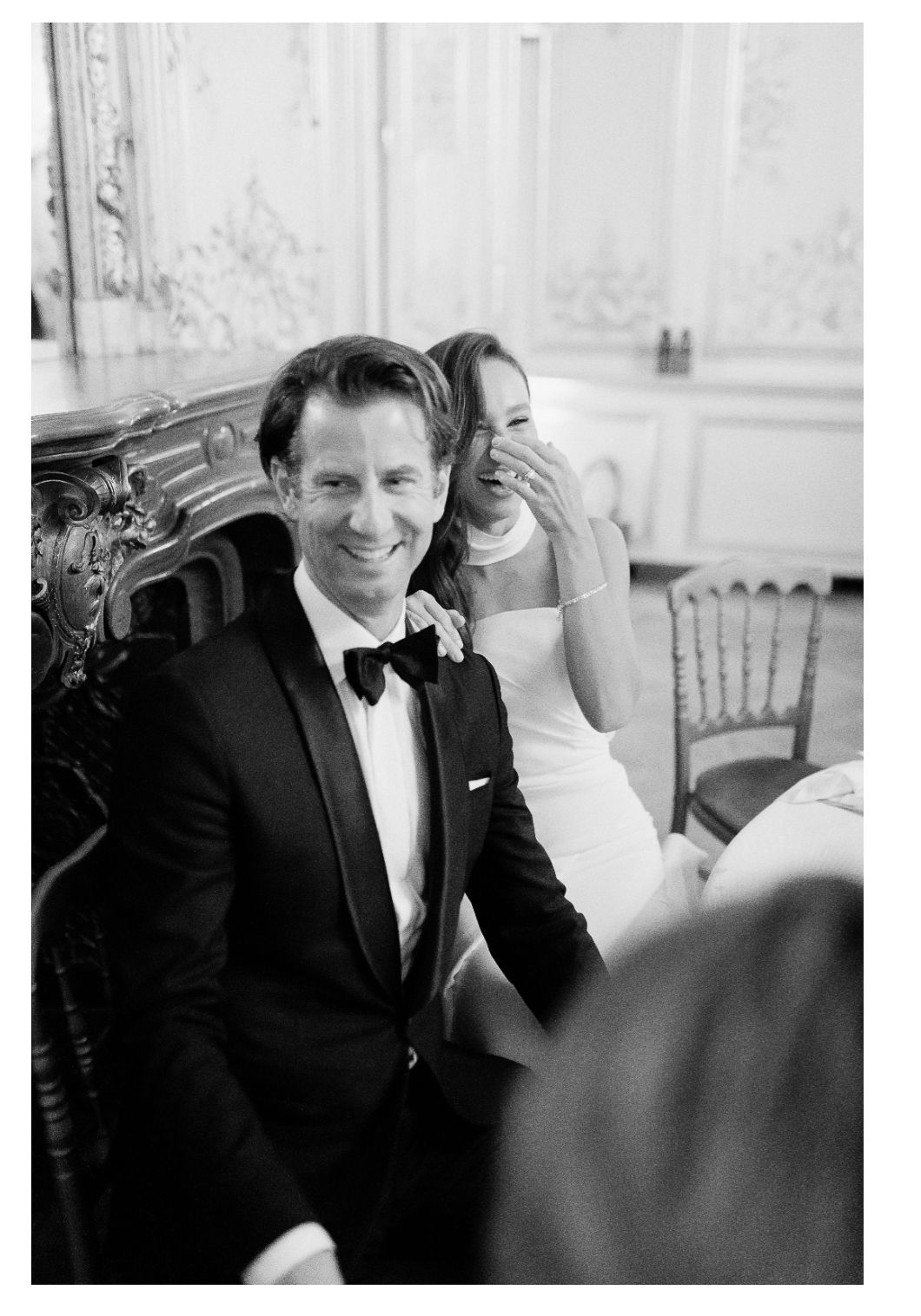 Black and white photo of a wedding celebration showing a candid moment at an elegant venue with ornate decor.