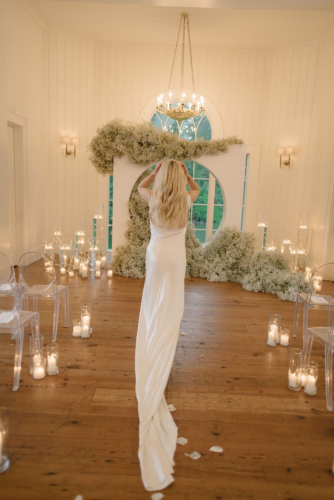 Bride in white gown stands before floral arch with candles and ghost chairs in elegant wedding ceremony venue.