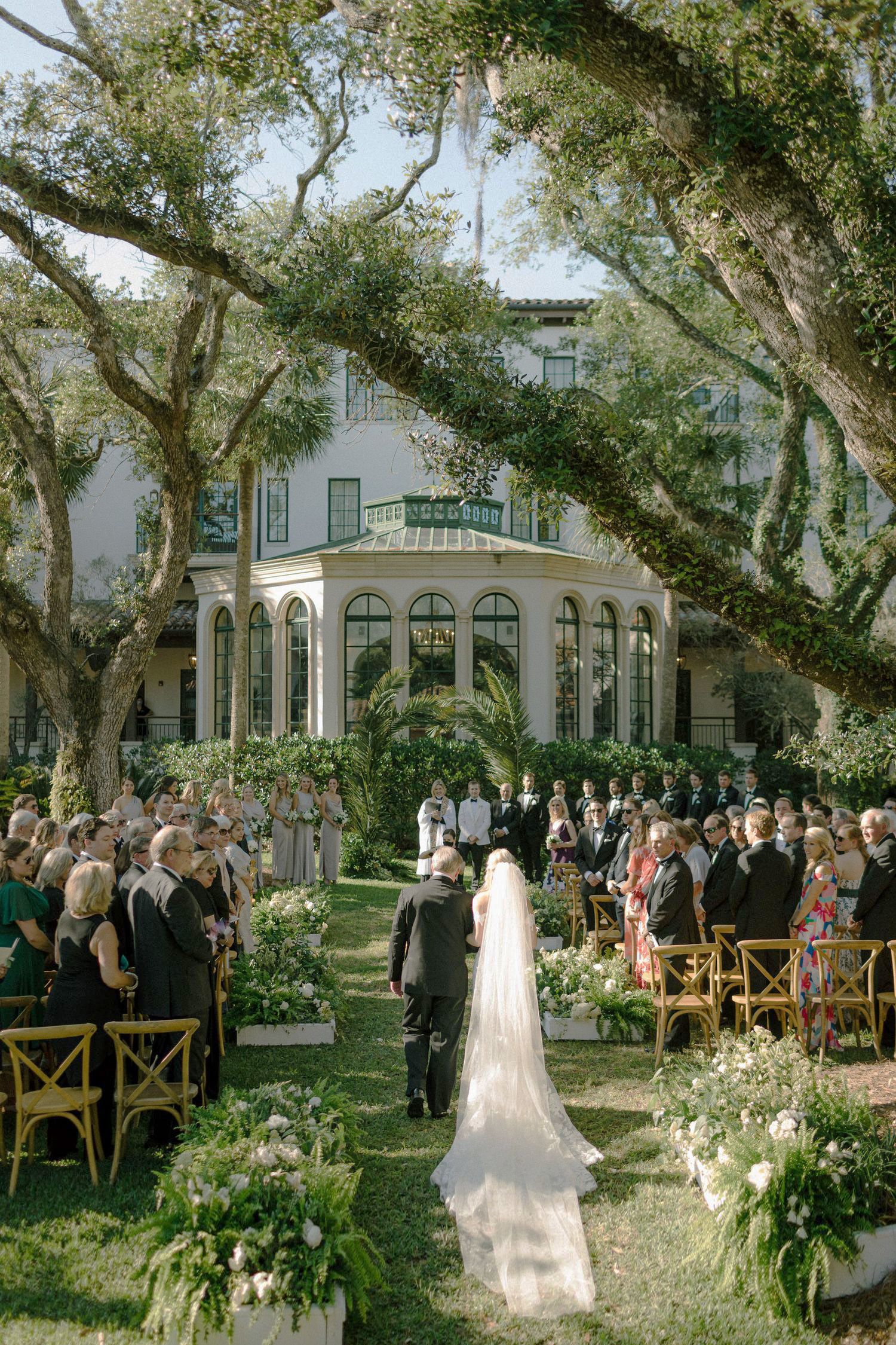 Bride walks down outdoor aisle at elegant garden wedding ceremony beneath oak trees at a white estate mansion.