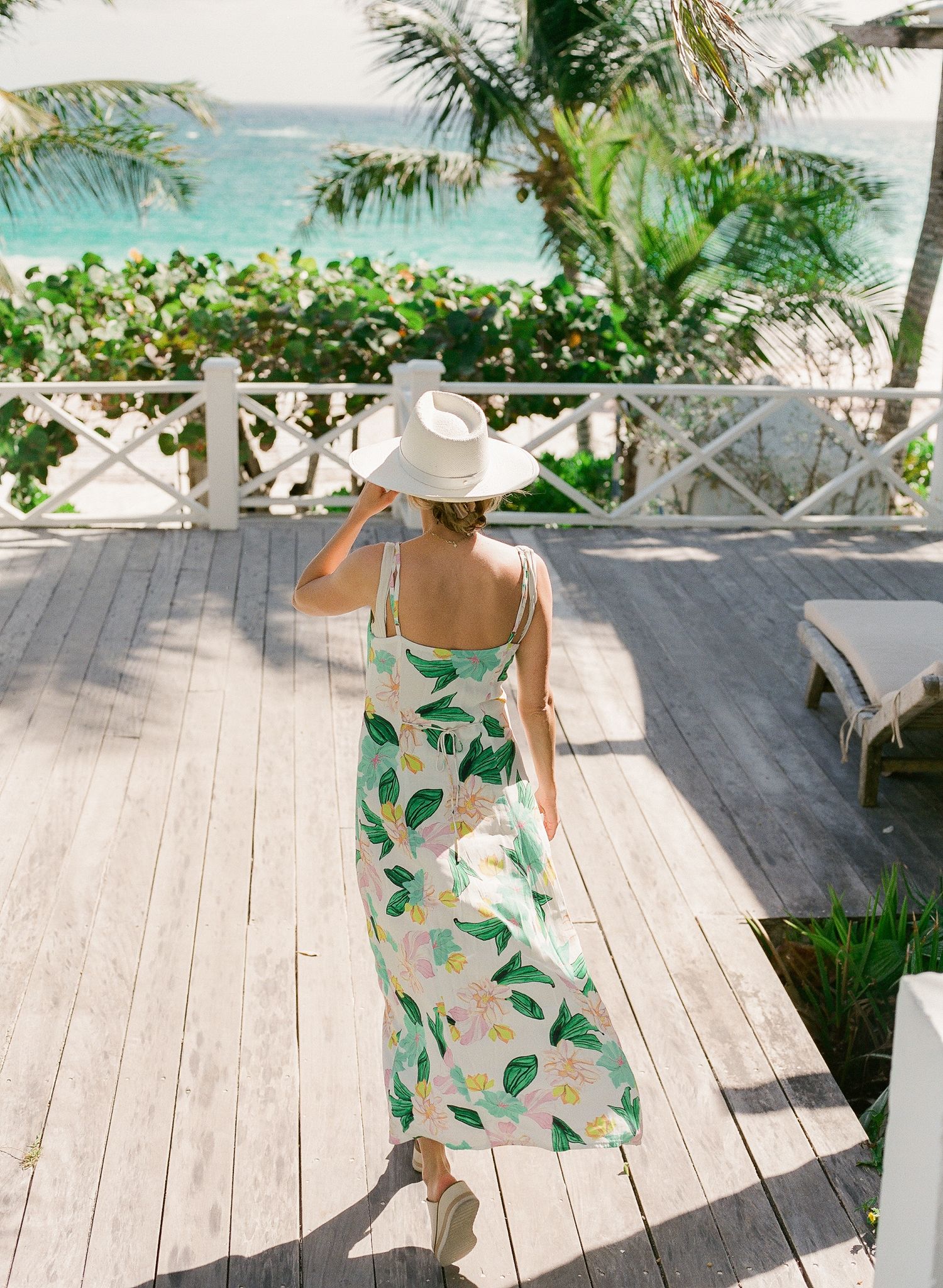 Woman in floral sundress and wide-brim hat stands on oceanfront deck with palm trees and turquoise waters in view.