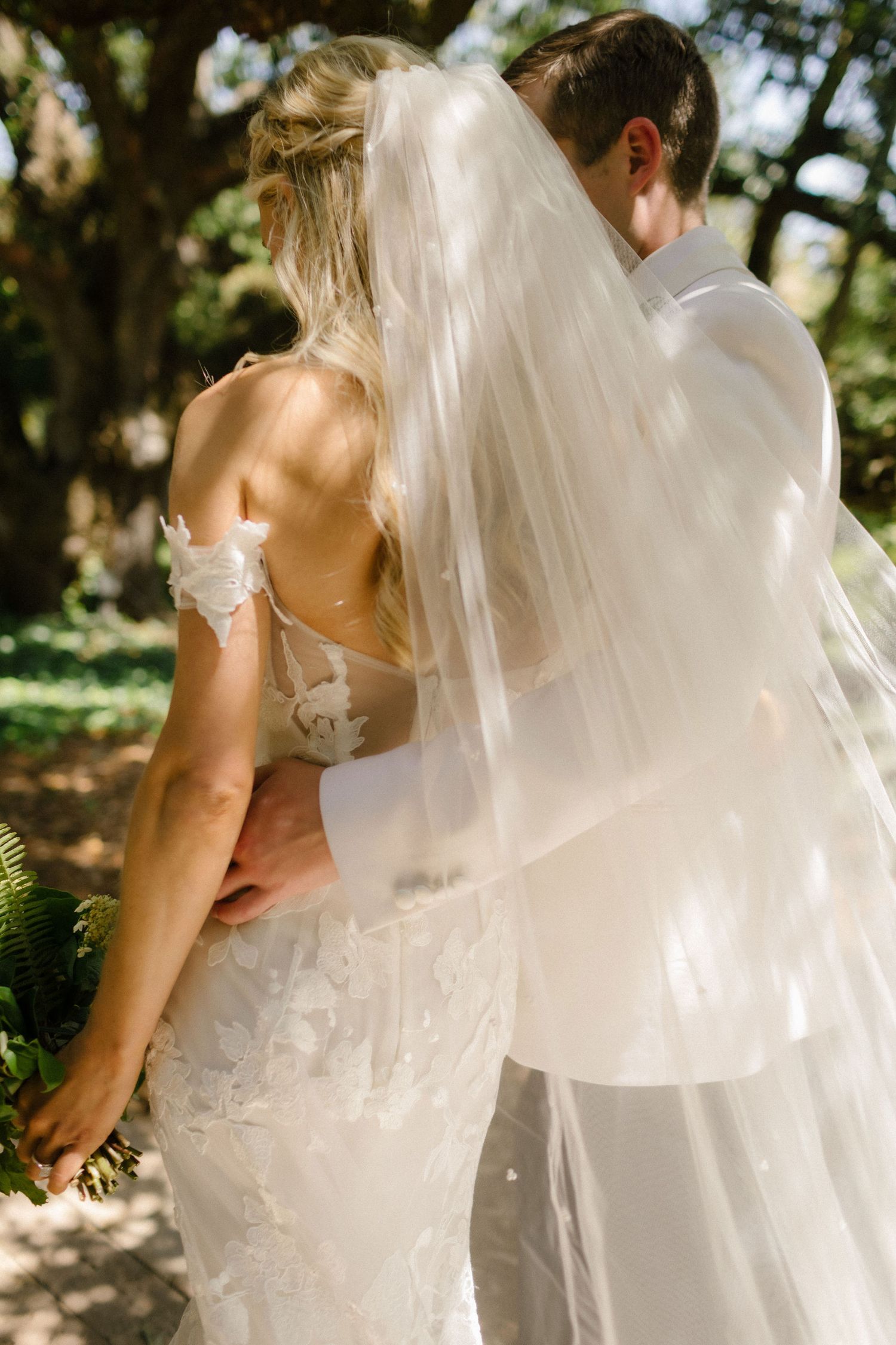 Bride and groom embrace outdoors, her lace wedding dress and flowing veil catching sunlight in a garden setting.