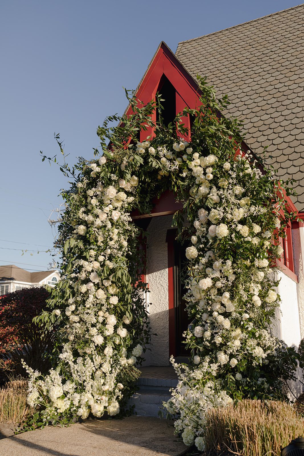 A stunning floral arch of white blooms and greenery frames the red door of a charming church entrance.