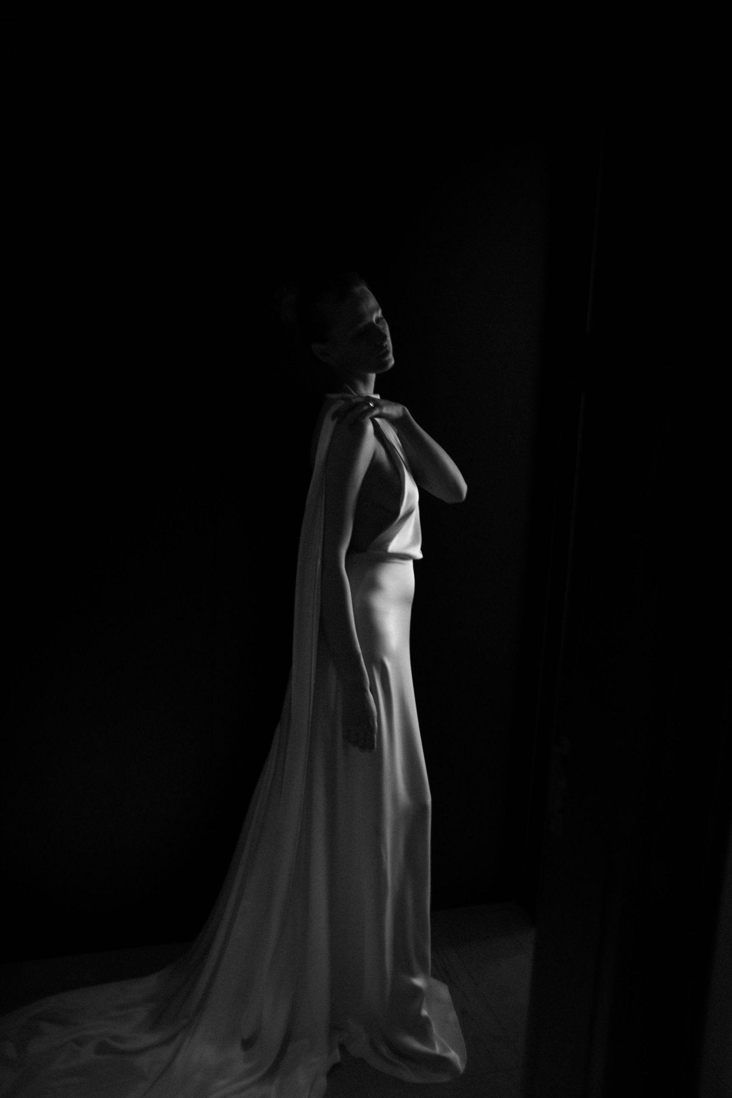 Elegant bride in silky white gown with dramatic cape train poses in dramatic black and white portrait.