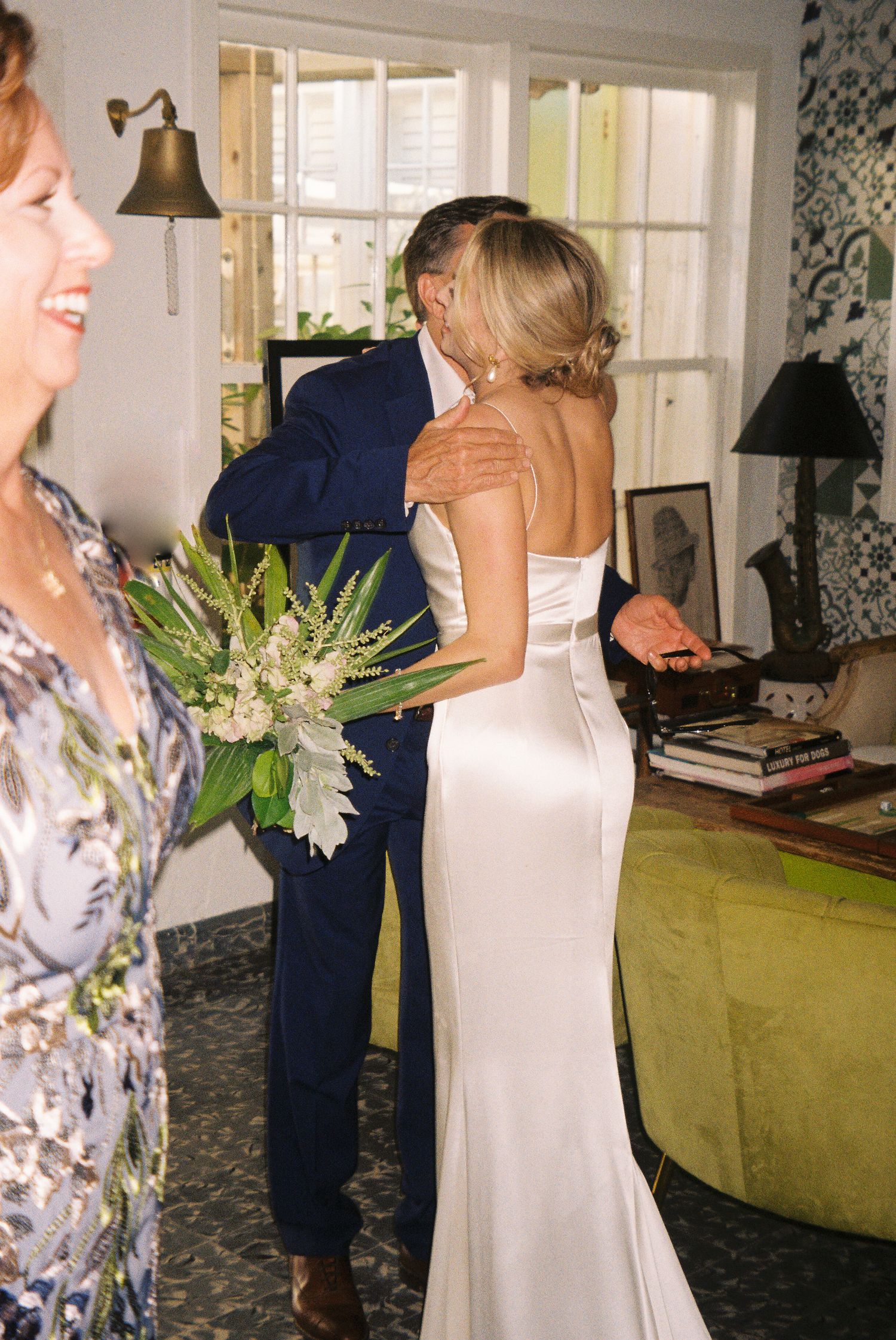 A bride in a fitted white gown holds a bouquet at an intimate indoor wedding gathering near a window.