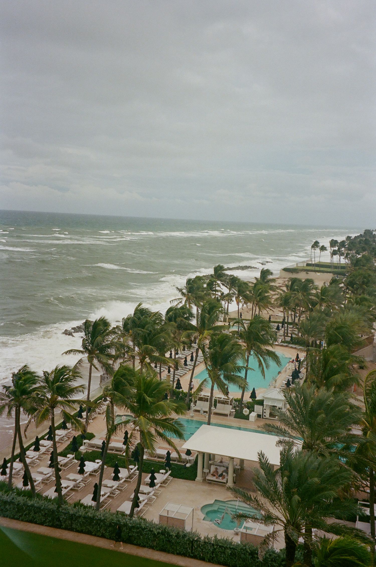 Aerial view of a beachfront resort with palm trees swaying in stormy weather and multiple swimming pools near the ocean.