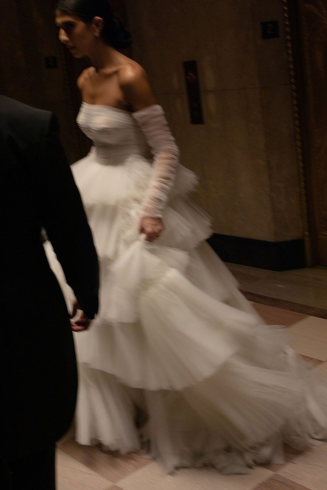 A bride in a flowing white strapless tulle gown with sheer gloves walks beside a man in a dark suit.