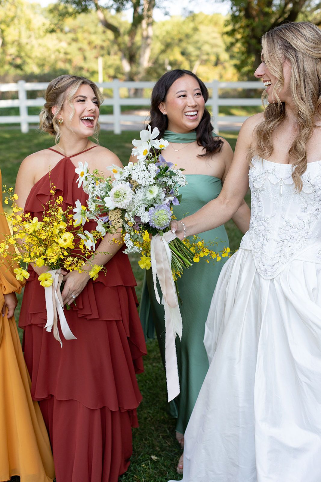 Wedding party members in colorful dresses sharing a joyful moment while holding floral bouquets outdoors.