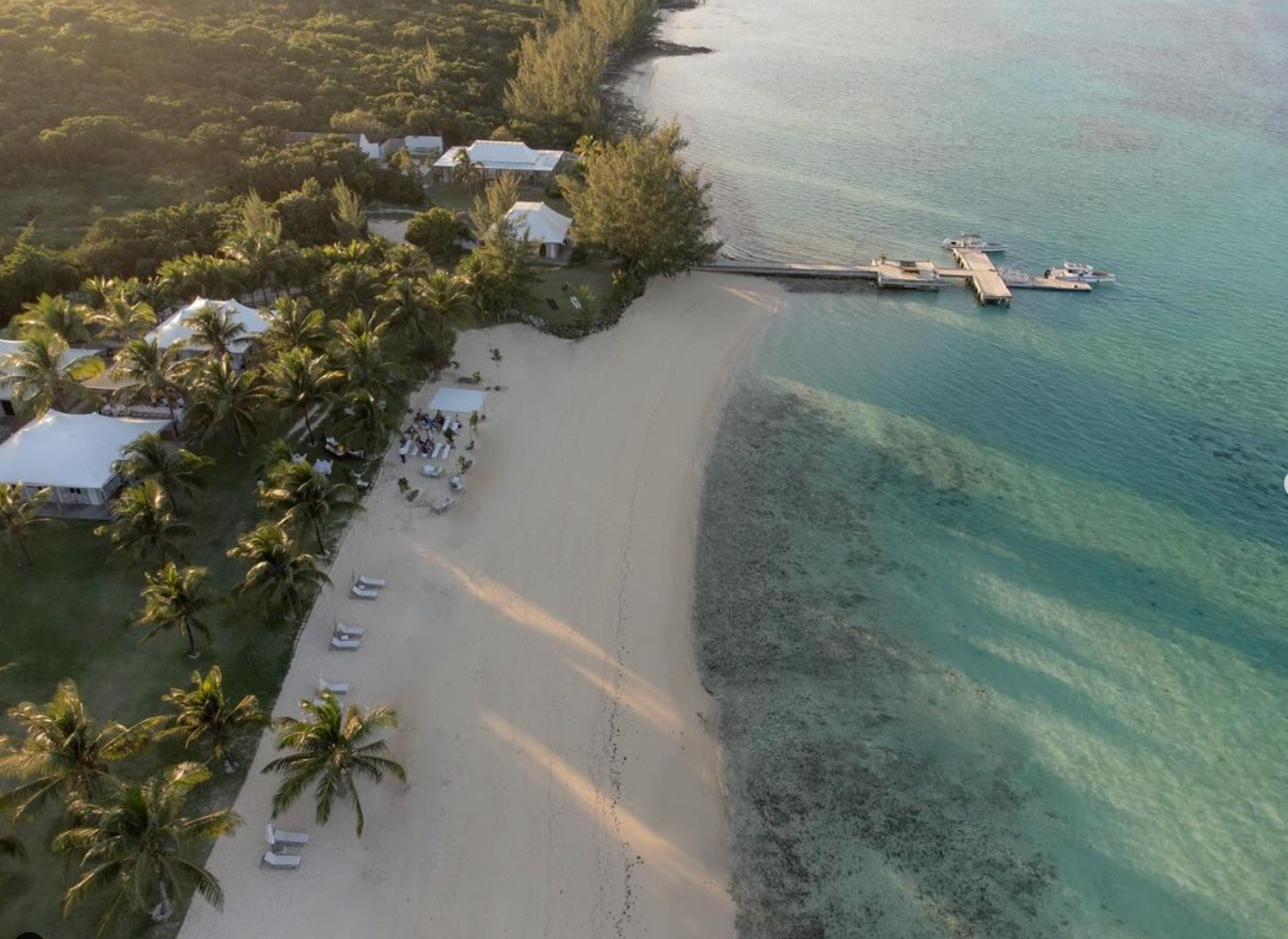 Aerial view of a tropical beachfront resort with wooden pier extending into turquoise waters and palm trees lining the shore.