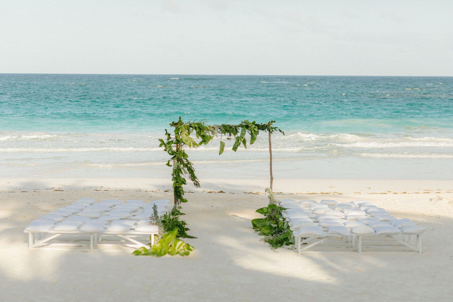 Beach wedding setup with a greenery arch and white benches on white sand with turquoise ocean backdrop.