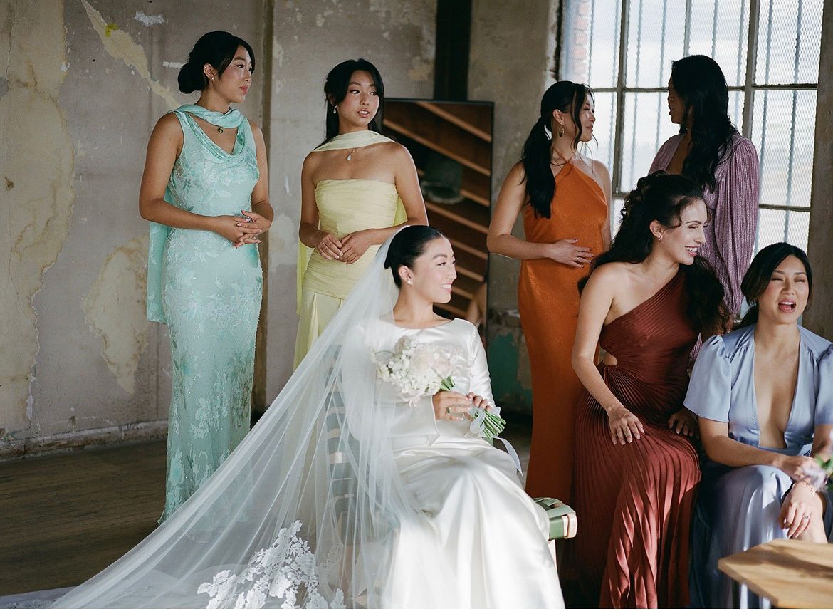 Bride in white gown with veil holds bouquet, surrounded by bridesmaids in colorful mismatched dresses in elegant venue.