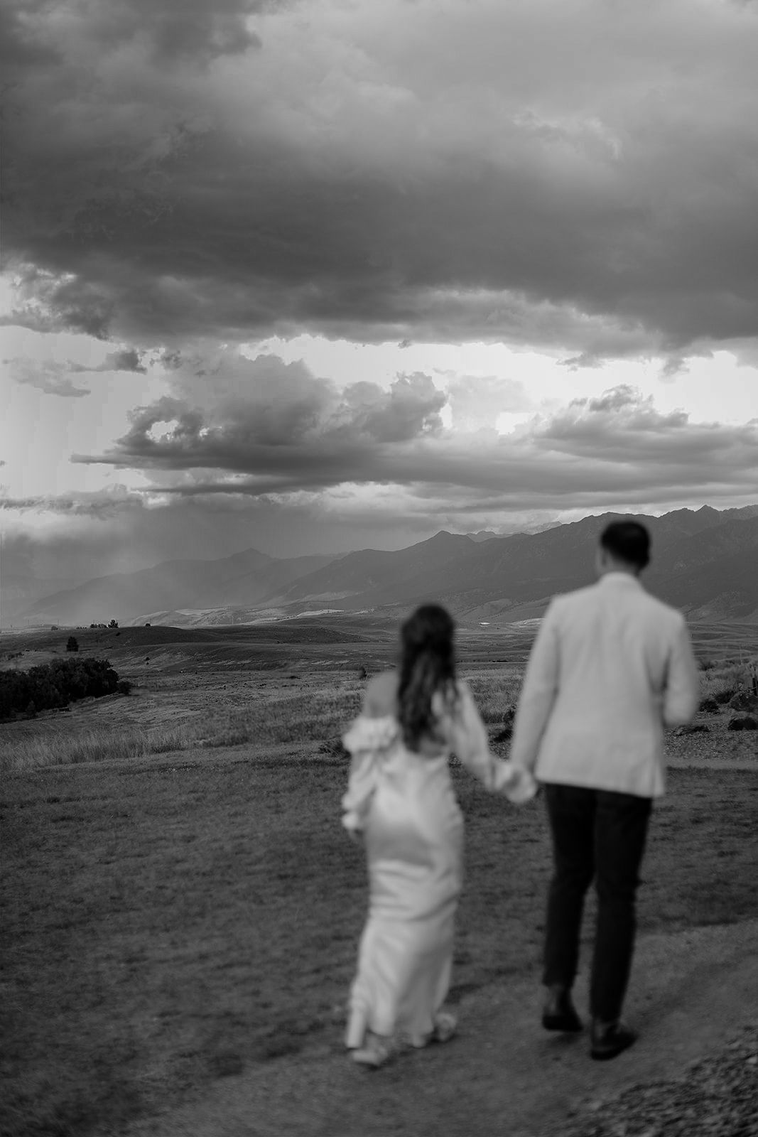 Black and white photo of a couple holding hands walking toward dramatic stormy skies over a vast mountain landscape.