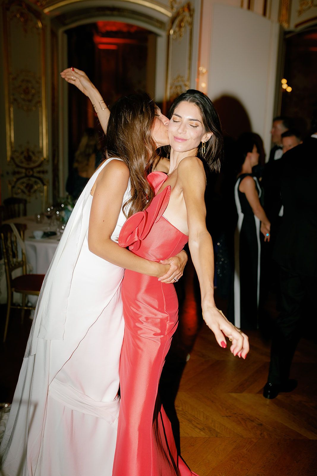 Two women in elegant gowns embrace and share a kiss at a formal event, one in white and one in red.