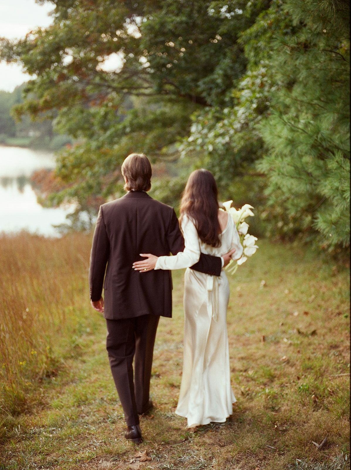 A couple in wedding attire walk together along a scenic lakeside path surrounded by lush greenery.