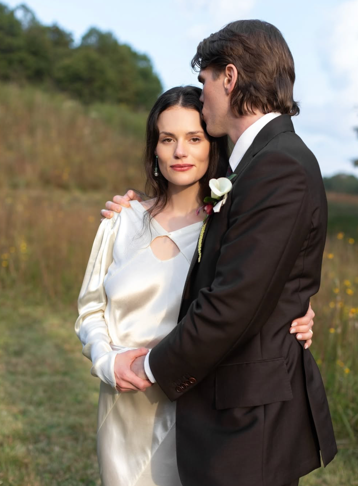 Bride in a satin ivory dress embraced by a groom in a dark suit with a white boutonniere in a sunlit meadow.