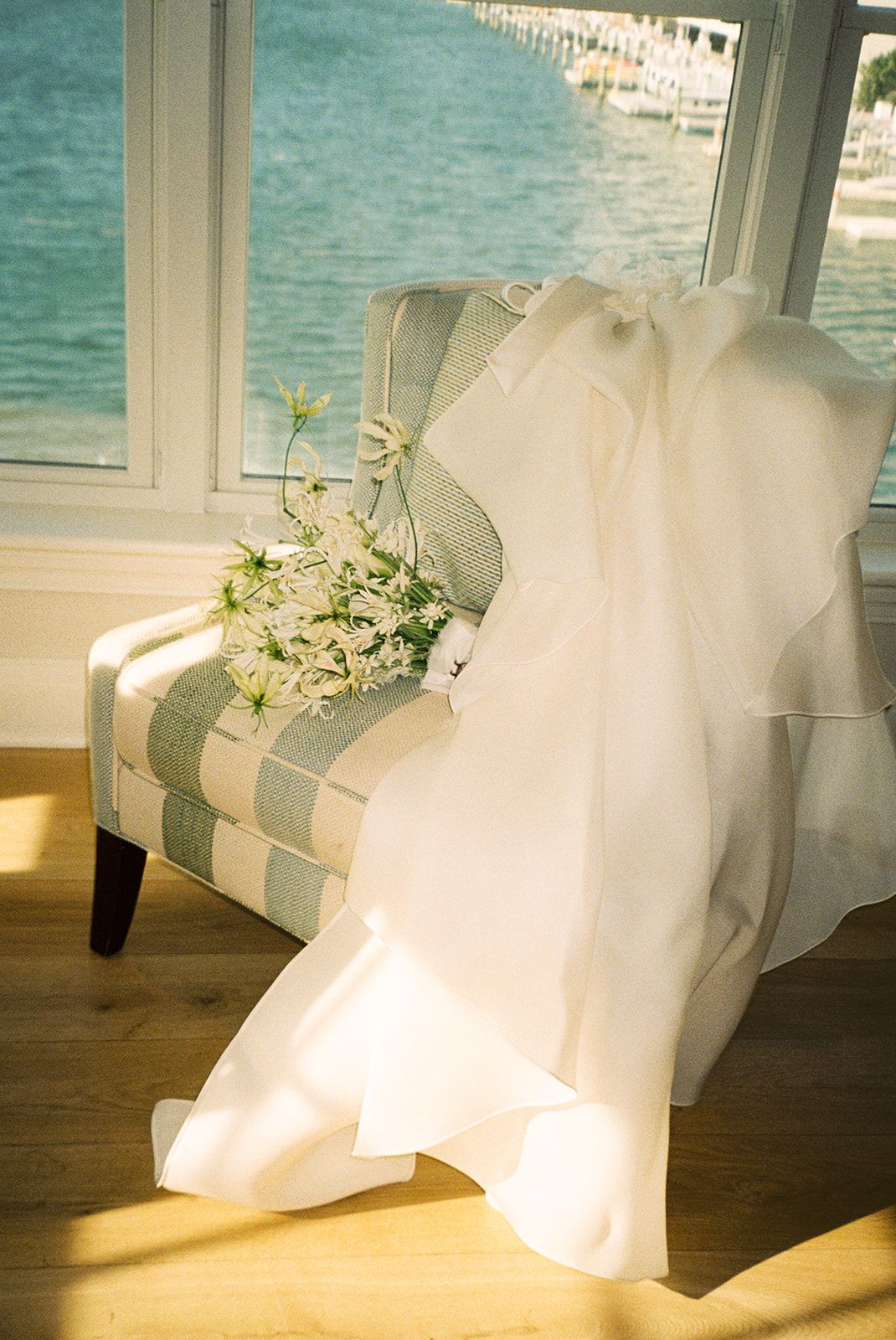 White wedding dress draped over striped armchair with floral bouquet, ocean view through sunlit window.