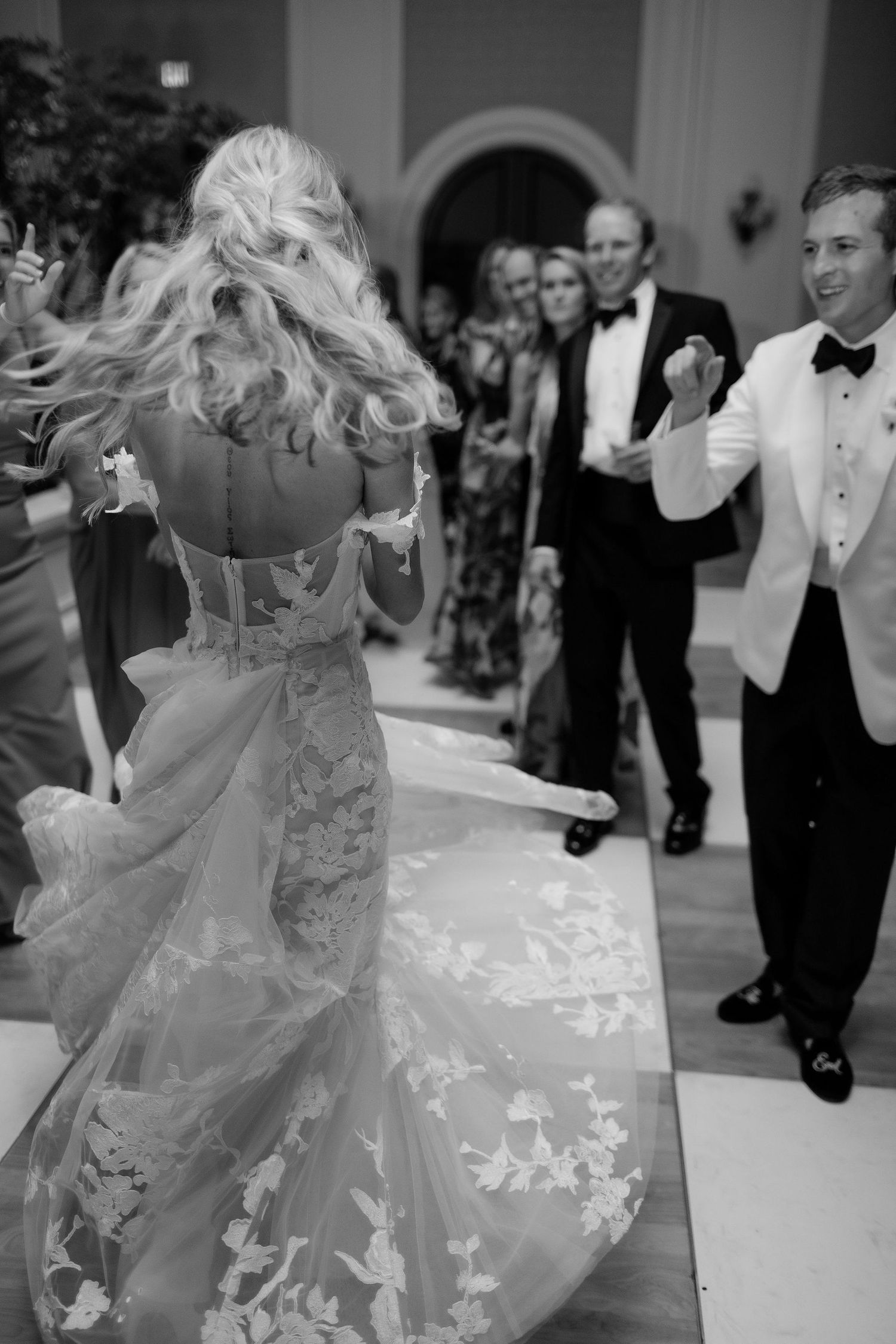 Bride twirls in floral lace wedding gown during first dance while groom in white tuxedo jacket smiles at reception.