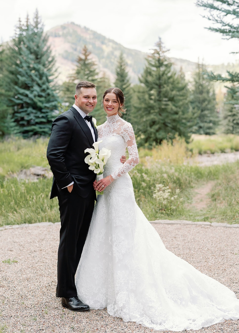 Bride in a white lace long-sleeve wedding gown and groom in a black tuxedo pose with a white bouquet among evergreen trees and mountains.