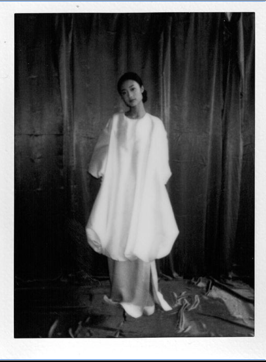Artistic black and white portrait with a person wearing a flowing white dress against a dark curtained background.