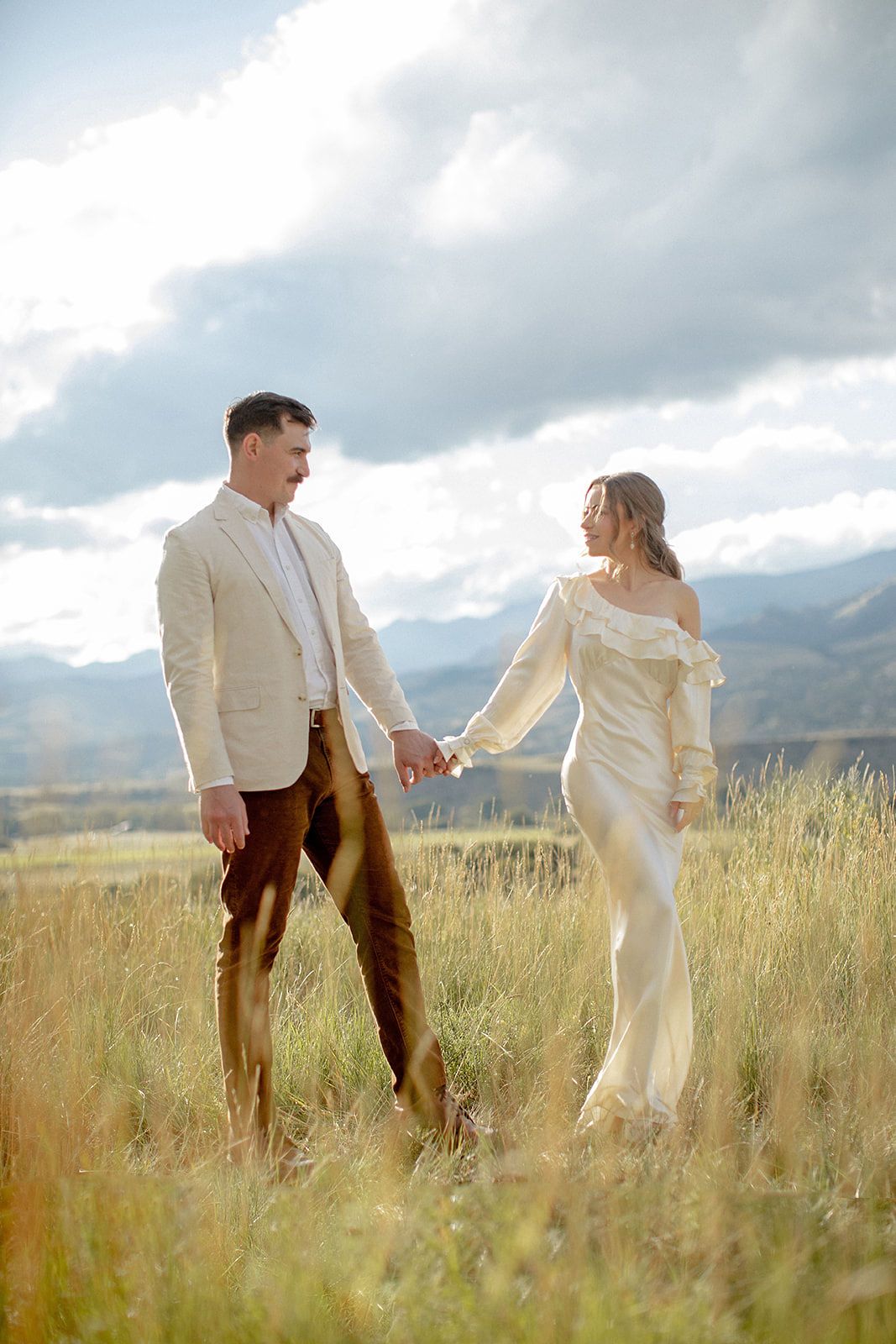 Couple in elegant wedding attire holding hands in a sunlit mountain meadow with dramatic cloudy skies.