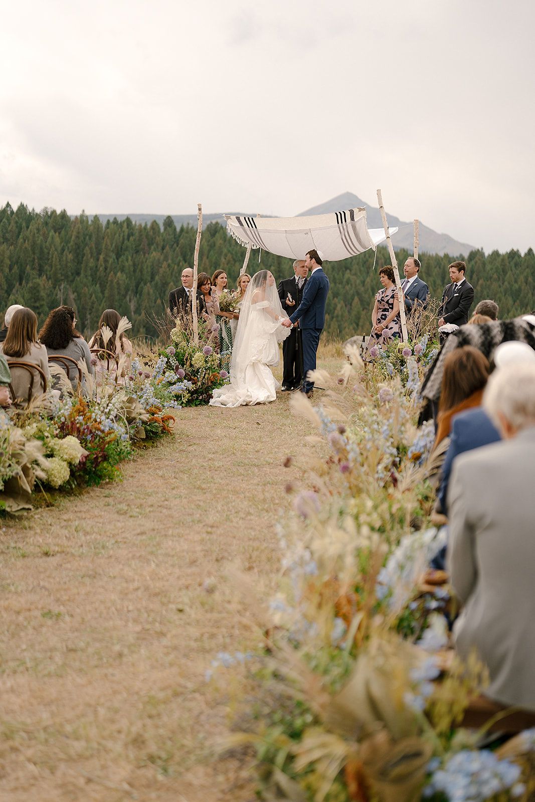 Outdoor mountain wedding ceremony with guests seated on both sides of a wildflower-lined aisle.