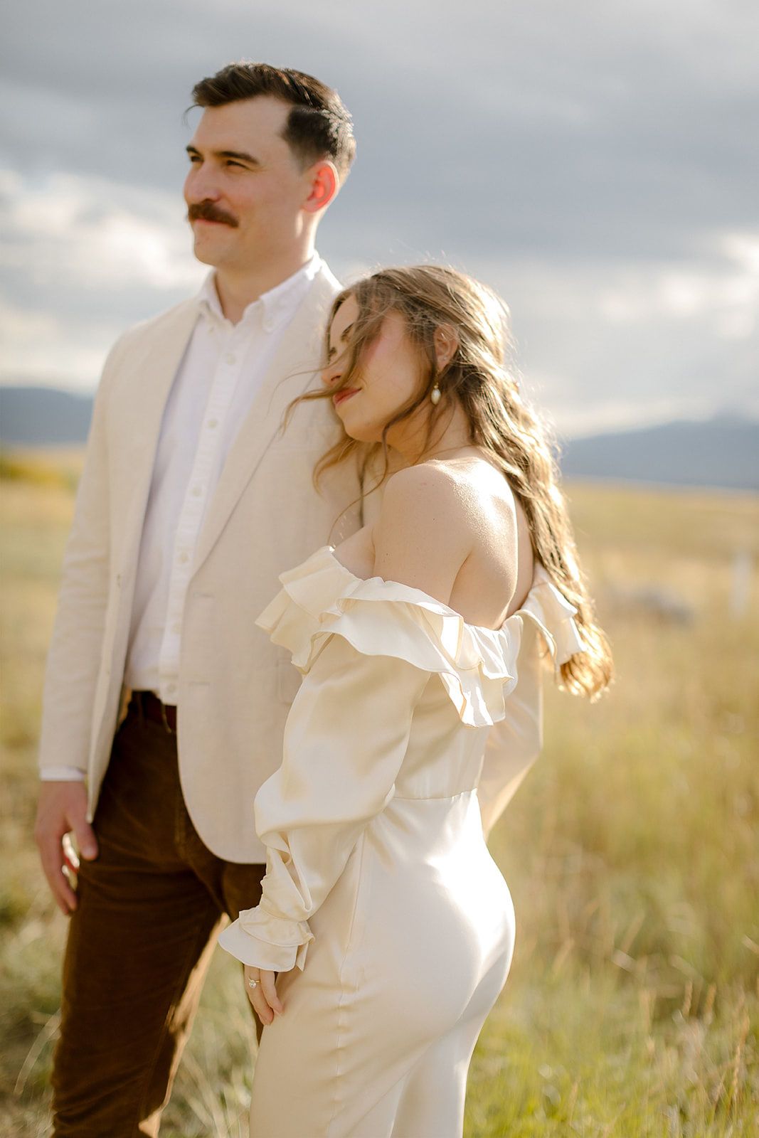 A couple in wedding attire embraces in a sunlit field with cloudy skies and golden grass in the background.