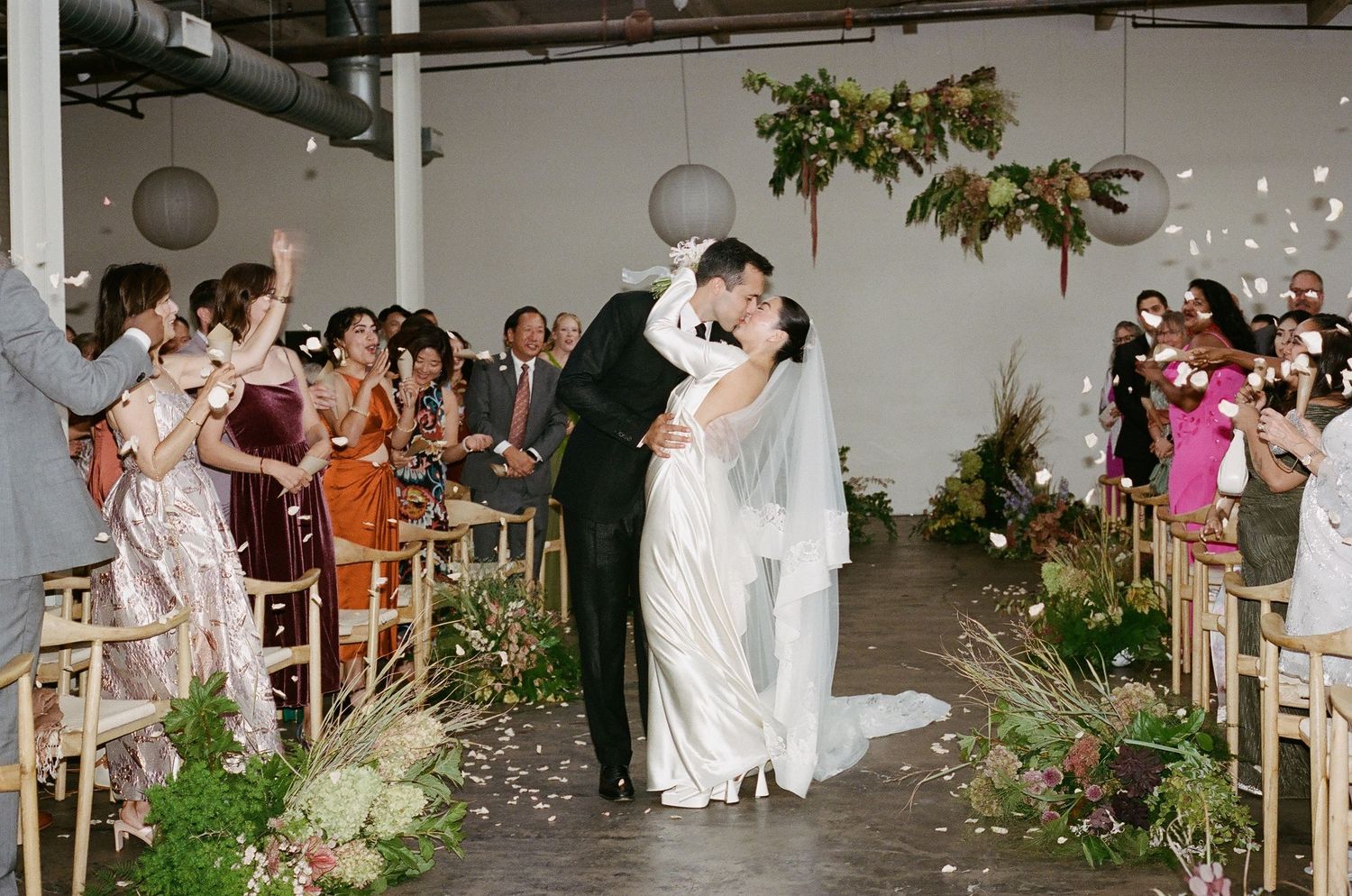 Wedding guests dance and celebrate on a rustic dance floor decorated with greenery and string lights at an indoor reception.