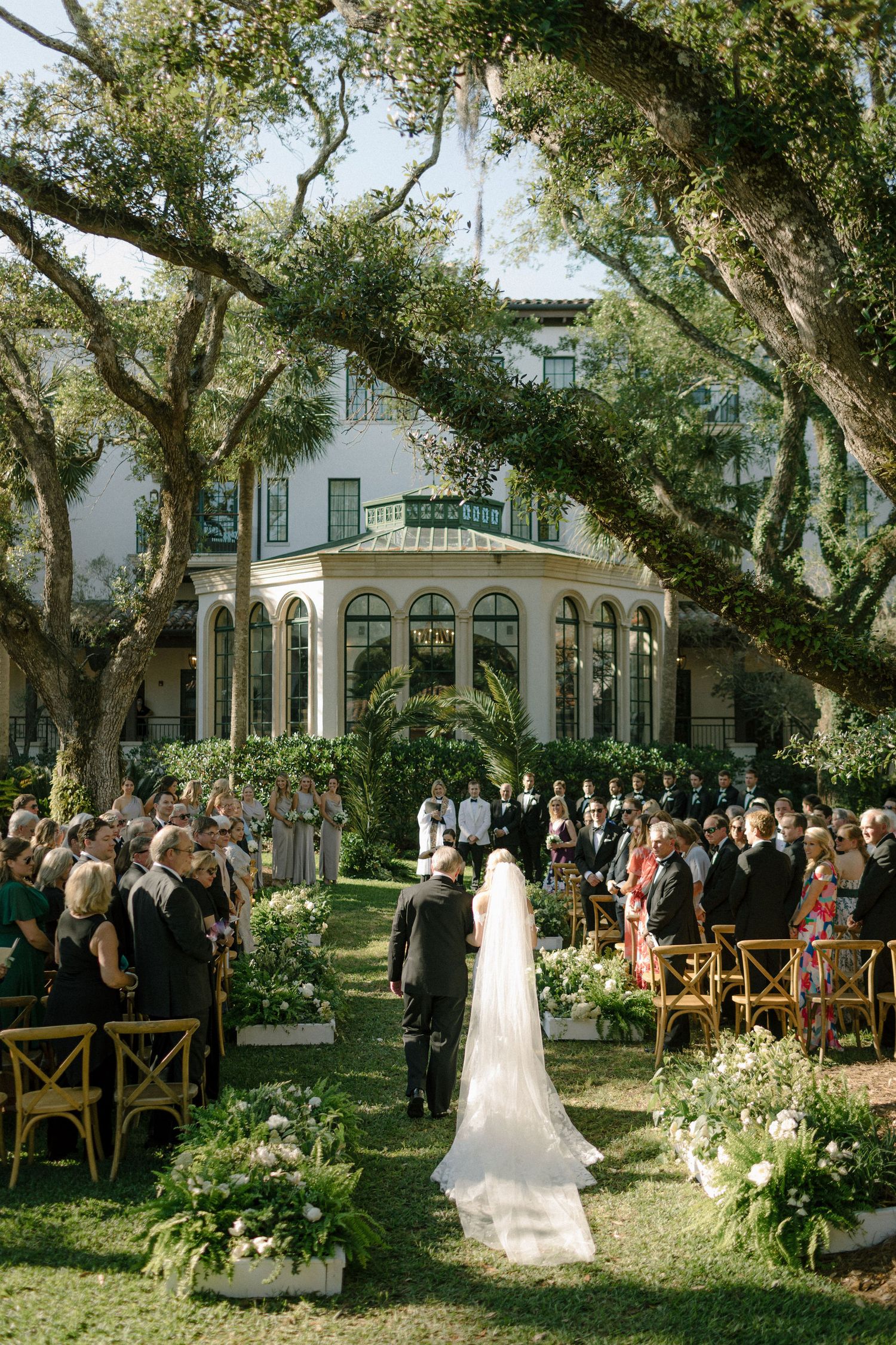 Bride walks down outdoor aisle at elegant estate wedding ceremony surrounded by lush trees and floral arrangements.