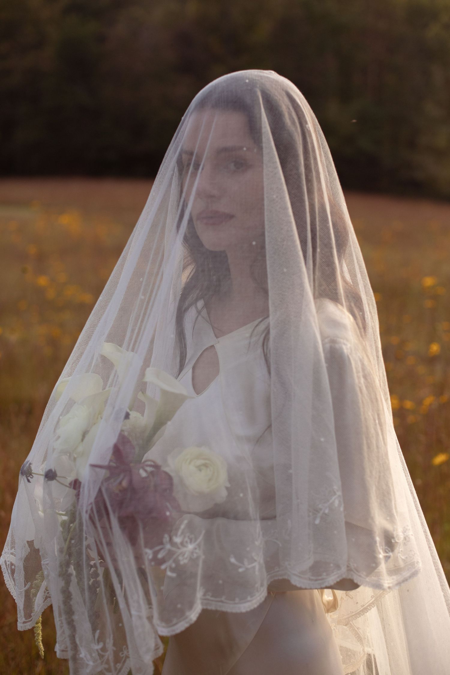 Bride in white wedding dress and lace veil holding bouquet of white roses in a sunlit field.