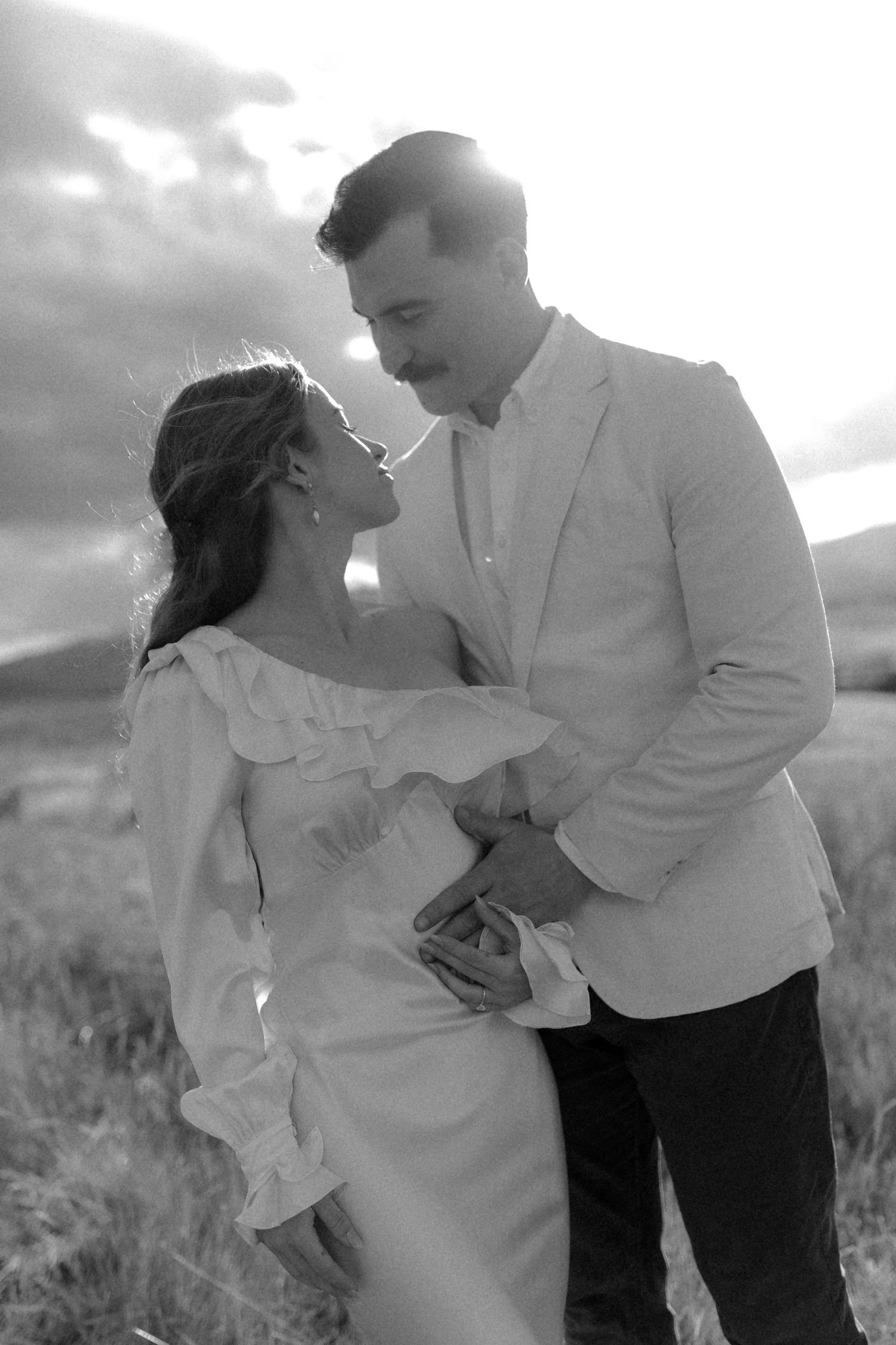 Black and white photo of a romantic couple embracing in a field at sunset, woman in ruffled dress, man in light blazer.