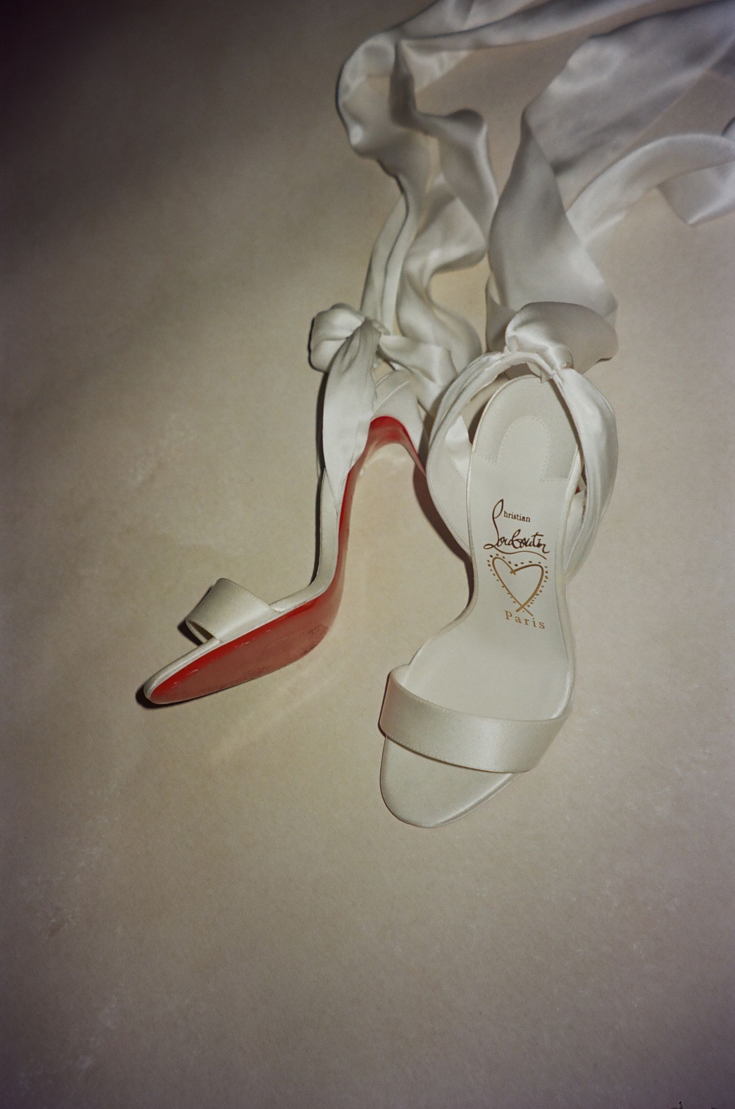 White Christian Louboutin heels with iconic red soles displayed next to a flowing white fabric.