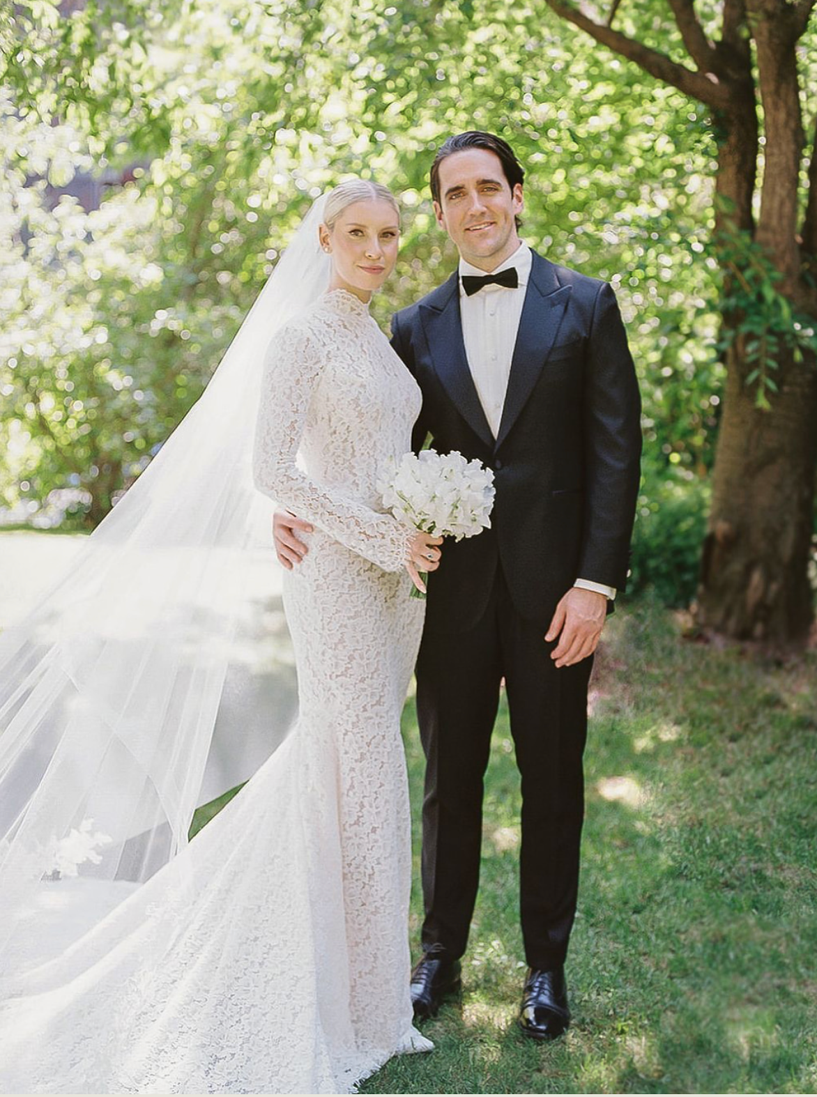 Bride in long-sleeve lace gown with veil and groom in black tuxedo posing outdoors among green trees.